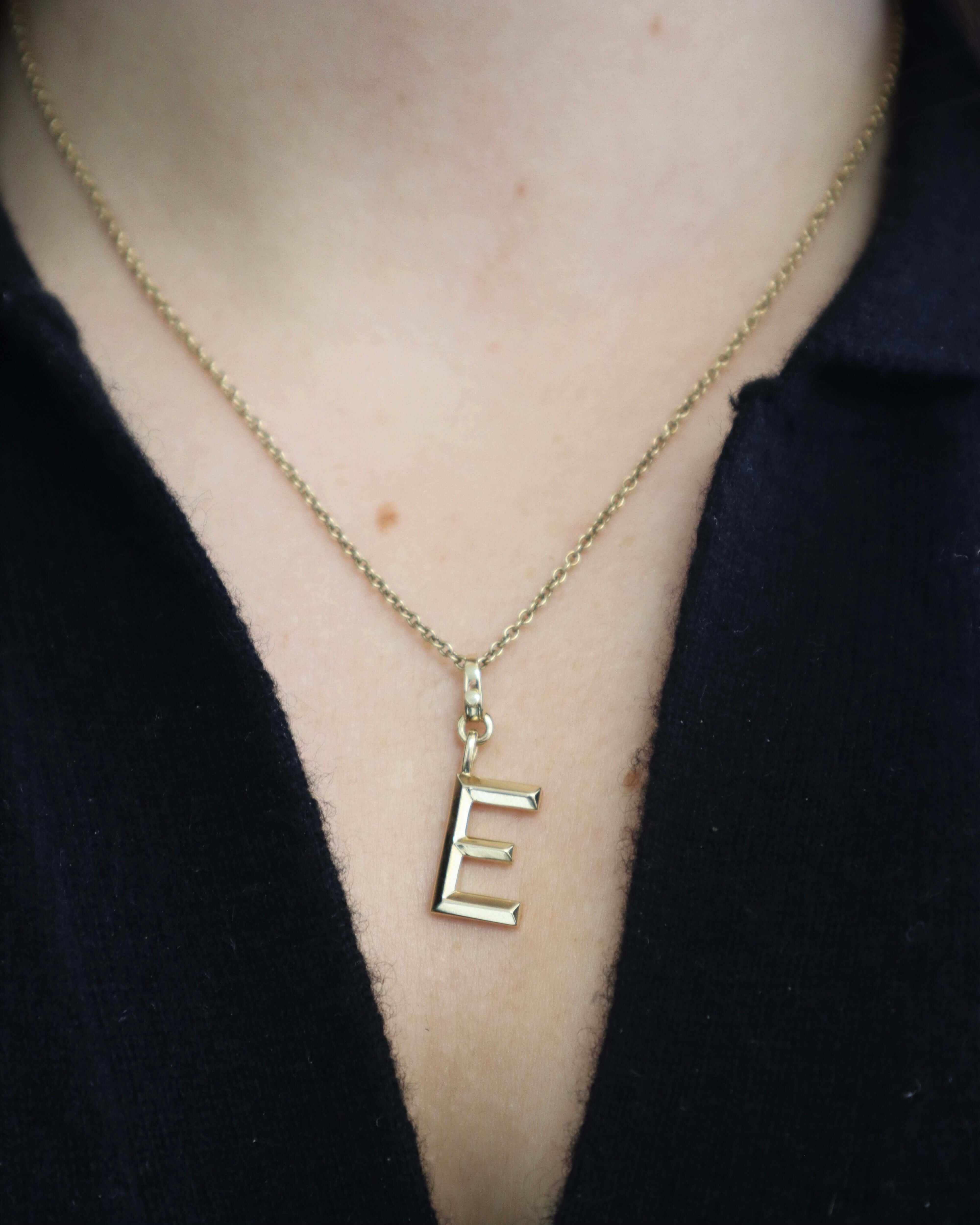 A close-up shows a person wearing a Janet Heller Fine Jewelry gold necklace with a Small Pyramid Initial Charm E, secured by a delicate clasp over dark clothing.