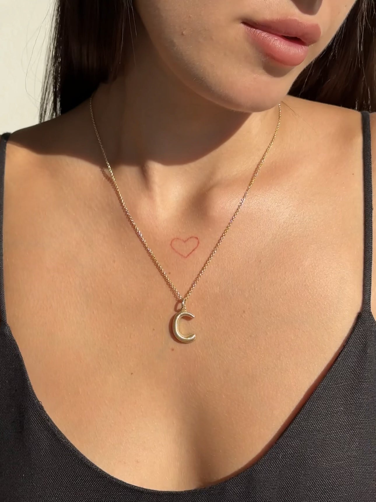 A woman in a thin black strap top showcases a Janet Heller Fine Jewelry Large Ridge Letter Charm gold necklace, along with a small red heart tattoo just below her collarbone.