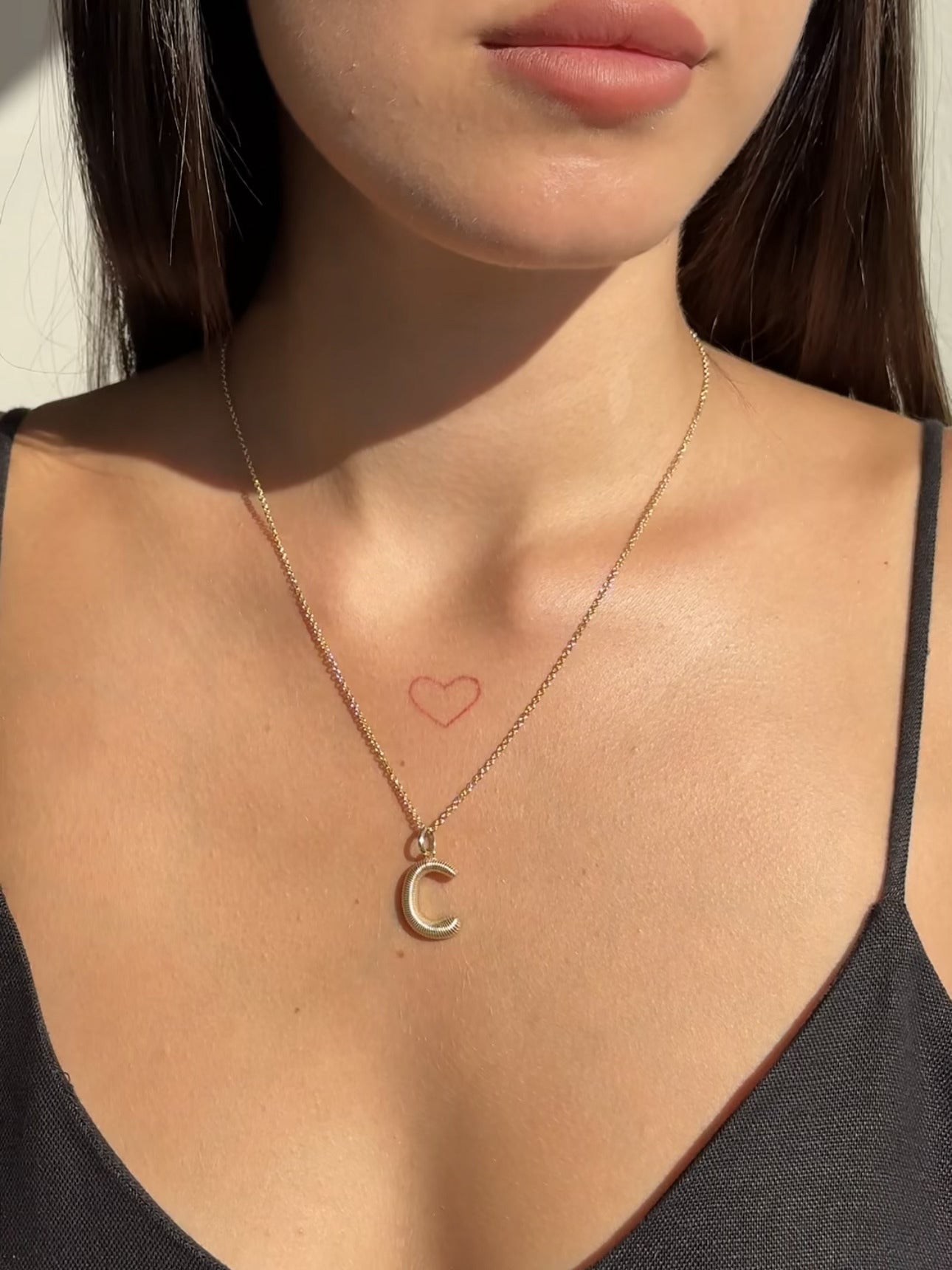A woman wearing a black top and a gold Janet Heller Fine Jewelry Large Ridge Letter Charm. She has long brown hair, and a small red heart outline tattoo on her upper chest adds custom charm.