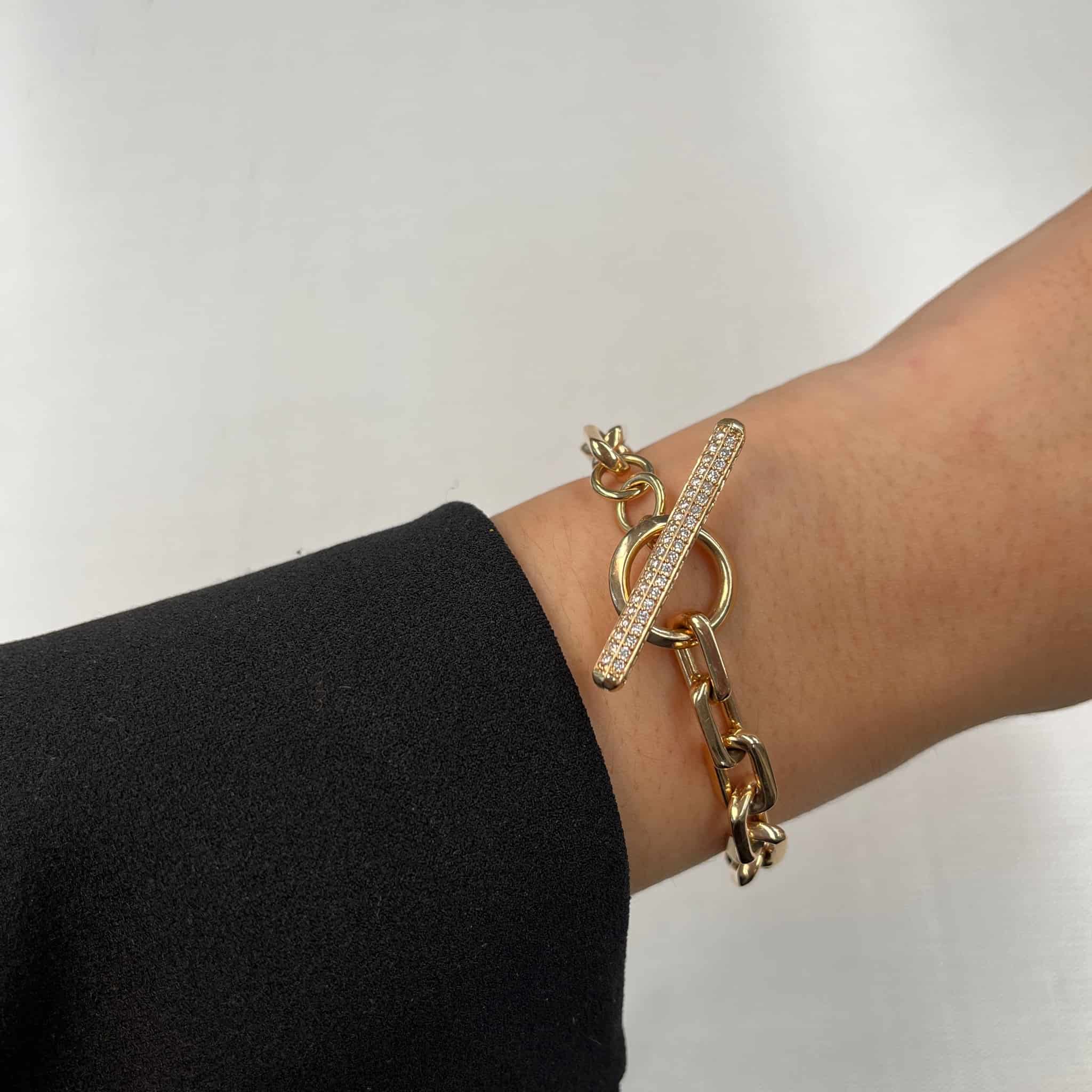 A persons wrist elegantly displays a Full Square Diamond Toggle Link Bracelet, boasting a gold chain and toggle clasp adorned with crystals by Janet Heller Fine Jewelry. Dressed in a black sleeve, they pose against a plain light grey surface.