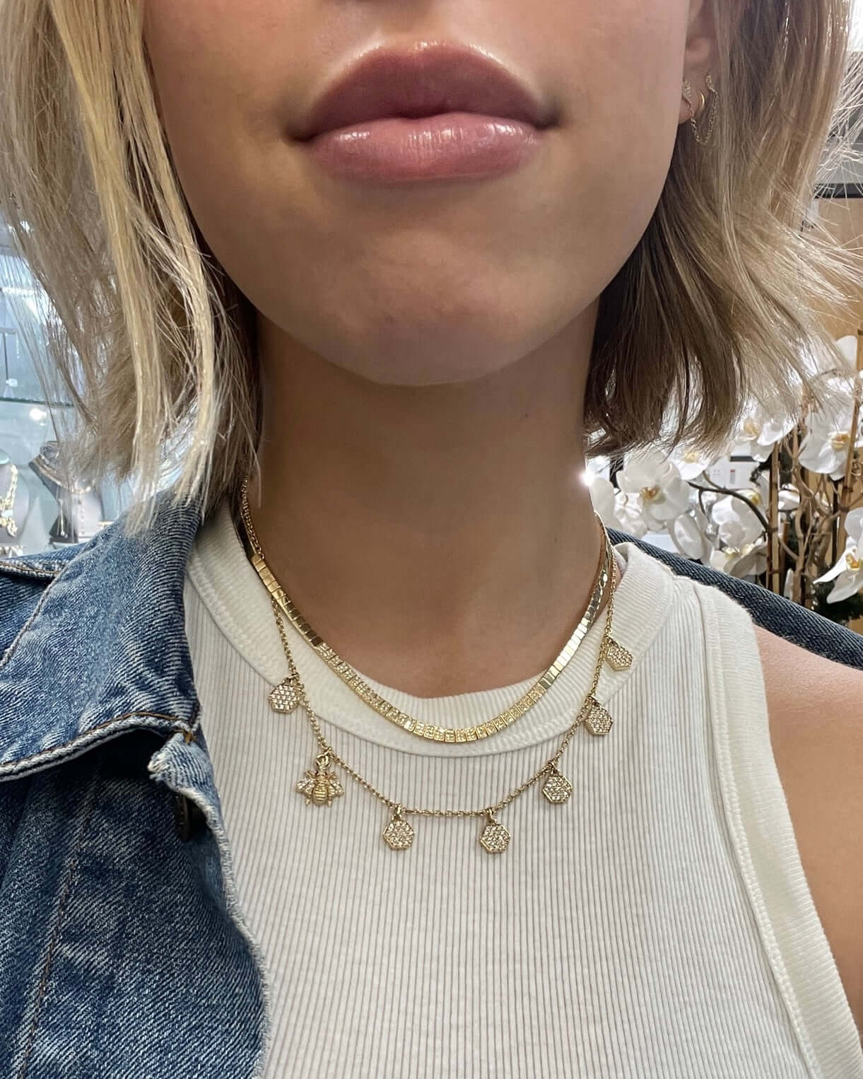 A person with short, wavy blond hair wears a Janet Heller Fine Jewelry Honeycomb & Bee Necklace over a white ribbed top and denim jacket, against a background of blurred white flowers.