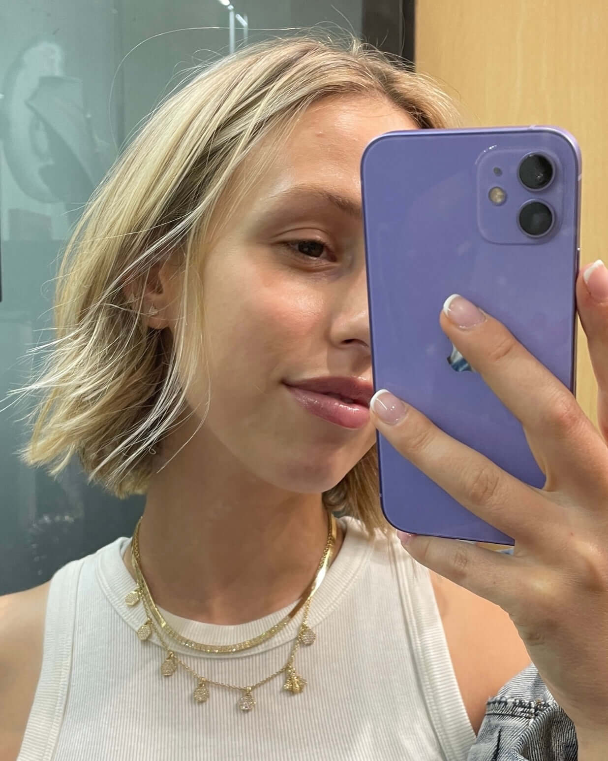 A person with short blonde hair takes a selfie using a lavender-colored smartphone, showcasing their white top and the Honeycomb & Bee Necklace from Janet Heller Fine Jewelry. Their soft light pink nails add a delicate touch to the stylish ensemble.