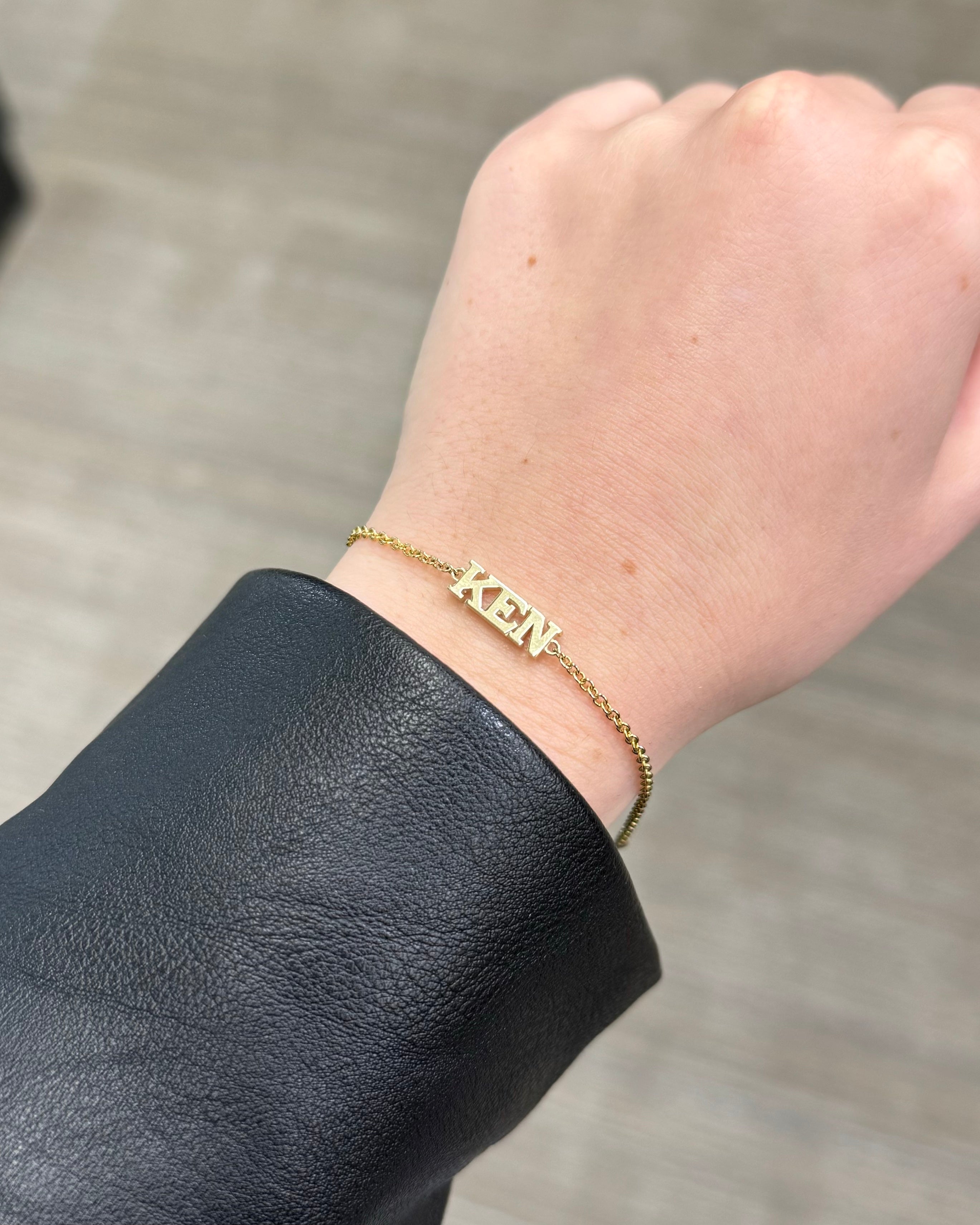 A wrist showcases the Block Name Bracelet, a gold chain adorned with ZEN in bold letters. The blurred neutral background enhances the bracelets personalized elegance, while the wearer’s black leather or faux leather sleeve adds a sophisticated touch to this unique accessory.
