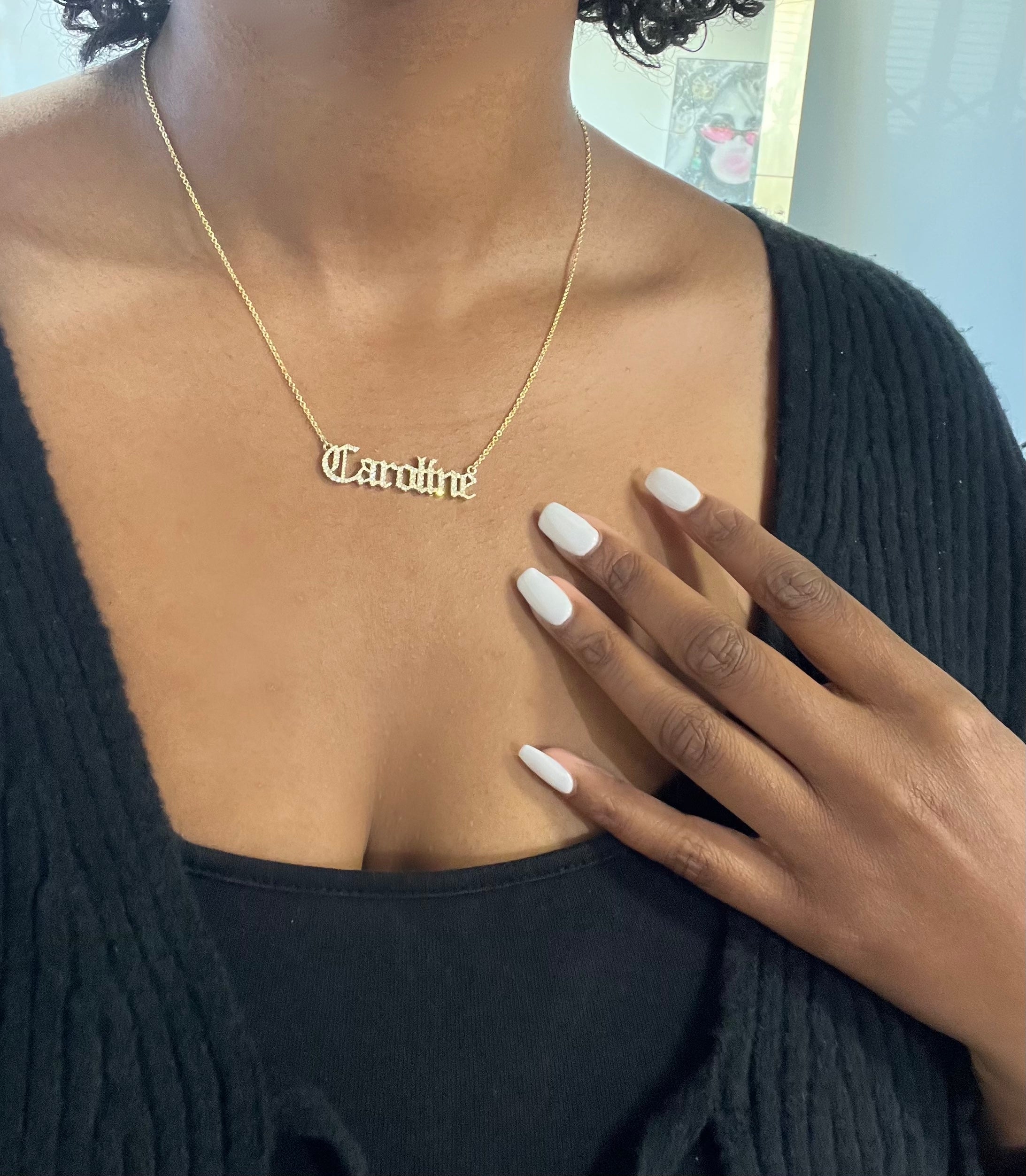 A person elegantly showcases their personalized jewelry—a Caroline Diamond Old English Name Necklace. Their manicured white nails rest gracefully on a black top paired with a cardigan, highlighting the unique piece.