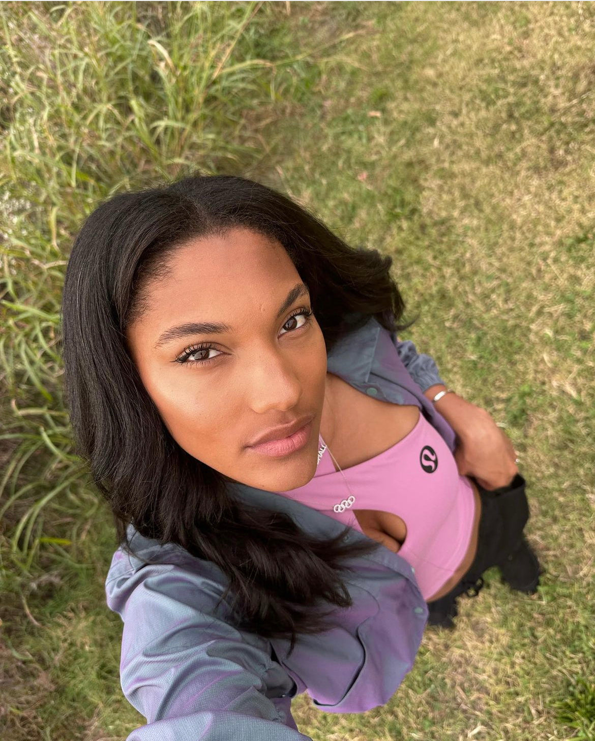 A woman with long dark hair takes an outdoor selfie from above, wearing a pastel purple top, light jacket, and the Diamond Rings Necklace—a perfect accessory for your final sale look—standing on grass with tall green plants behind her.