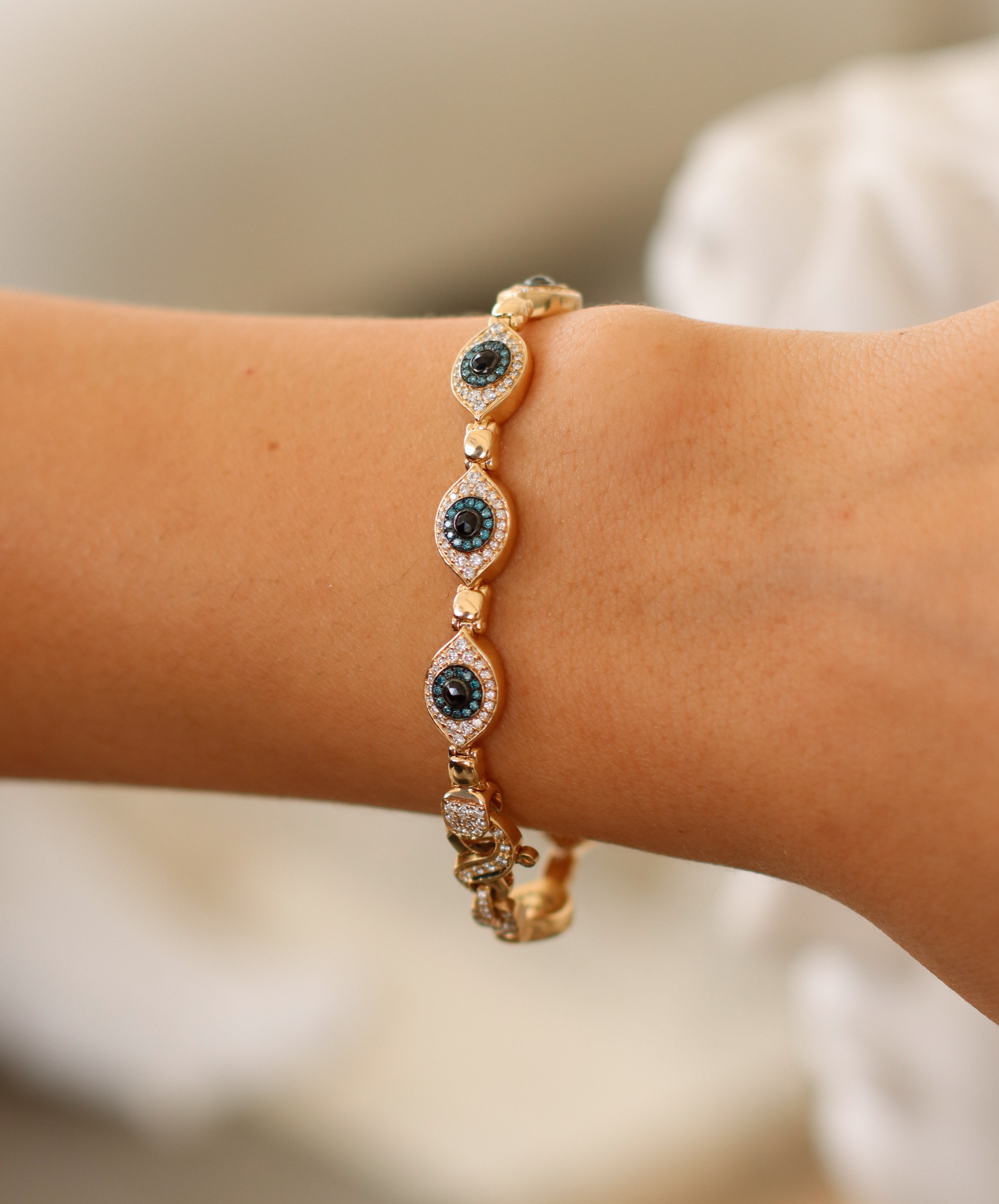 A close-up showcases a Repeating Evil Eye Bracelet from janethellerfinejewelry.com, featuring blue and white gemstones in an eye design. The intricate detailing contrasts against smooth skin with a blurred background, highlighting the exquisite customizations.