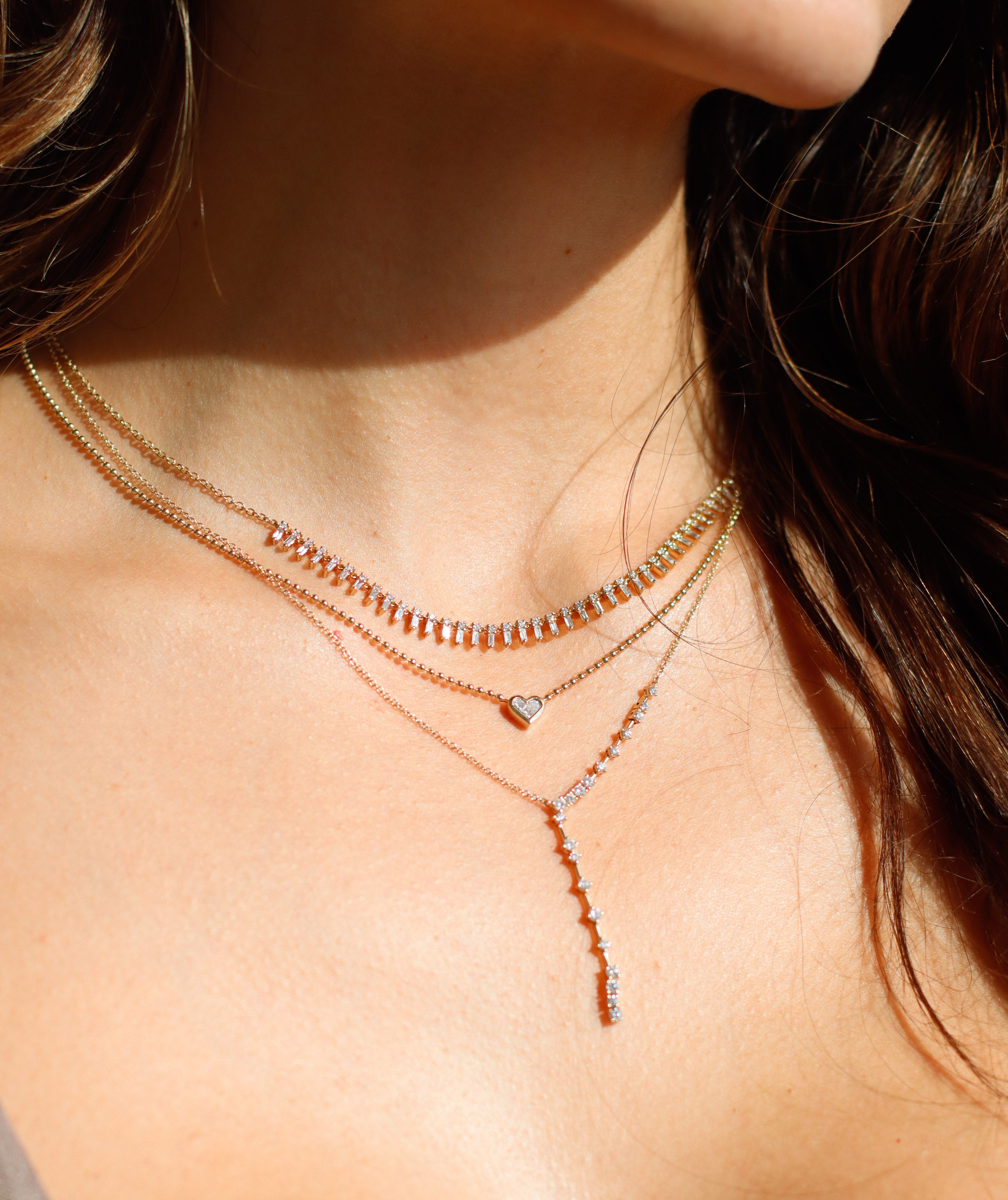 A person with long brown hair, wearing a sleeveless top, showcases three layered necklaces from Janet Heller Fine Jewelry: a simple chain, one with small stones, and the Diamond Constellation Lariat Necklace featuring a heart-shaped pendant and an extended line of stones.