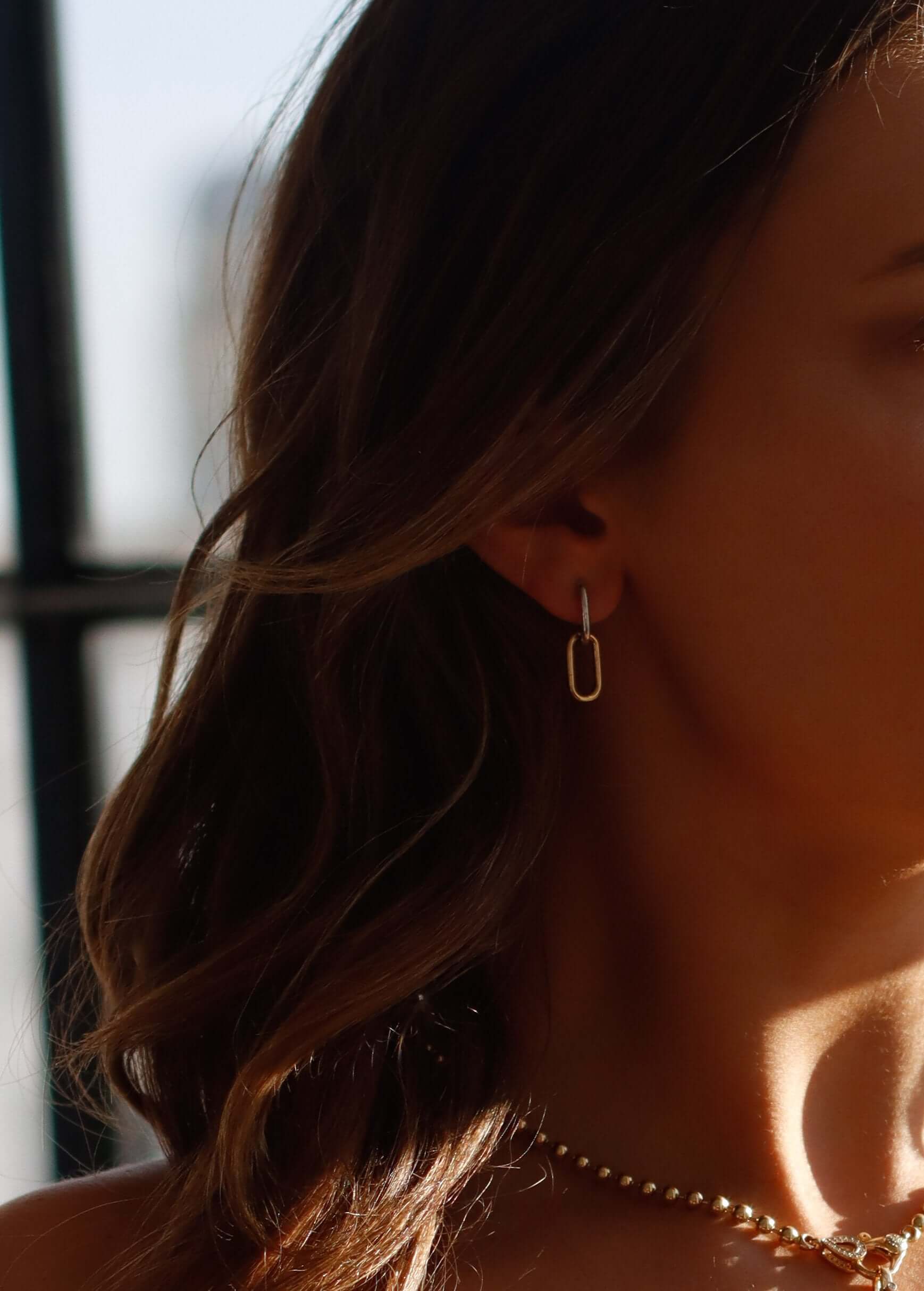 Close-up of a woman showcasing exquisite jewelry: Two Tone Oval Link Earrings paired with a delicate beaded necklace. The softly blurred background and sunlight cast a warm glow on her hair and skin, enhancing the elegance of these customizable pieces.