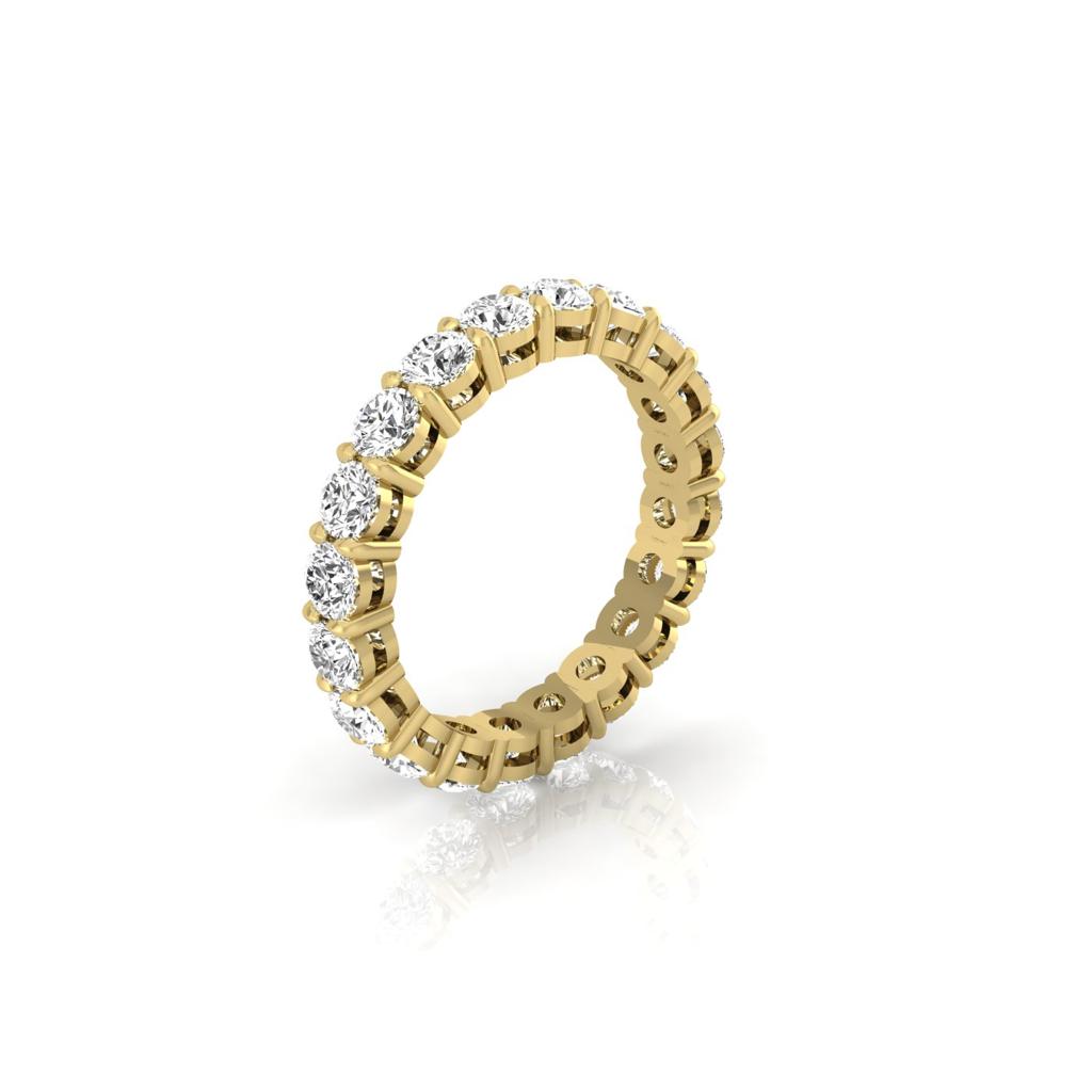 The Full Lab Grown Diamond Eternity Ring showcases 18KT gold with a continuous row of round, sparkling diamonds in a prong setting. Its F Color VS Clarity stones brilliantly catch the light, casting soft shadows on the white surface below.