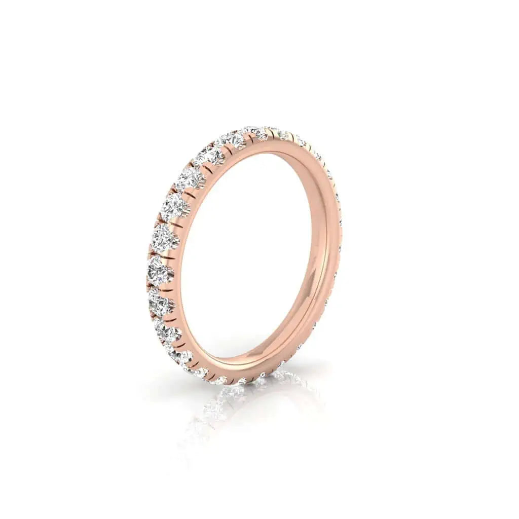 The 1ct Round Diamond Eternity Ring, crafted in 18KT rose gold, showcases small VS clarity round diamonds encircling the band. Its delicate elegance is highlighted on a white background with a reflective surface enhancing its sparkle.