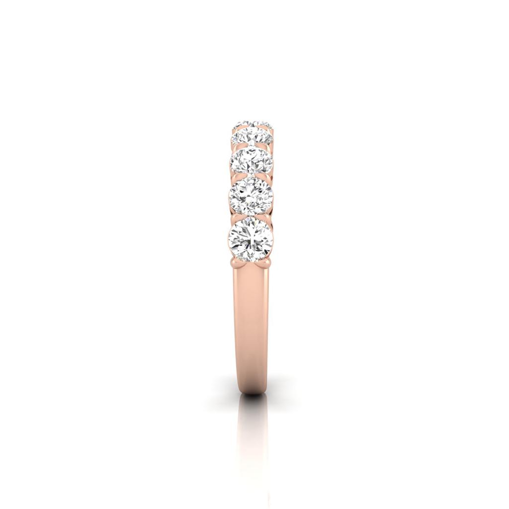 The Half Diamond Eternity Ring, designed in 18KT rose gold, features a row of seven dazzling round diamonds set in a straight line on top. Its displayed against a plain white backdrop, accentuating the diamond layout and warm gold hue.