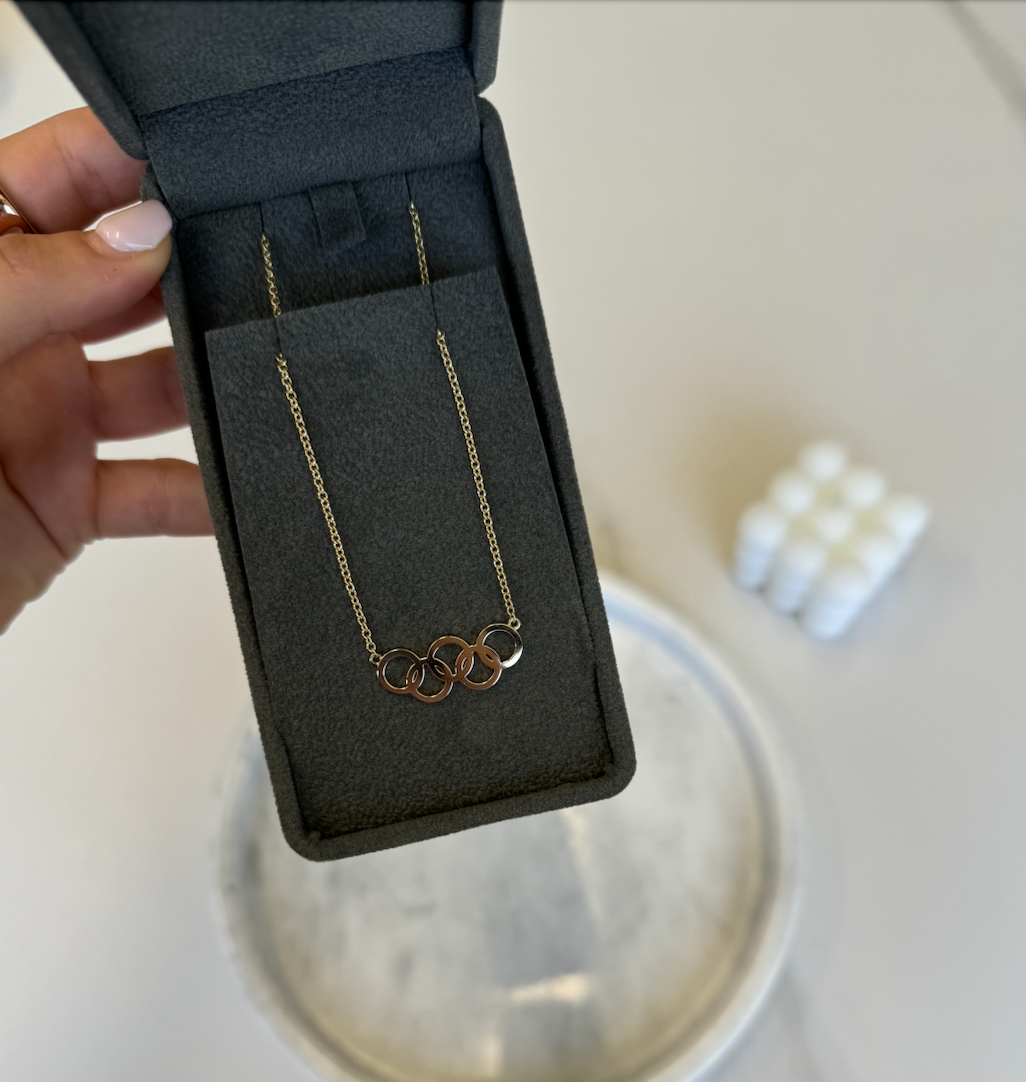 A hand holds an open black jewelry box displaying the Gold Rings Necklace, featuring three interlocking gold rings. In the blurred background, there is a round surface and a small stack of white objects.