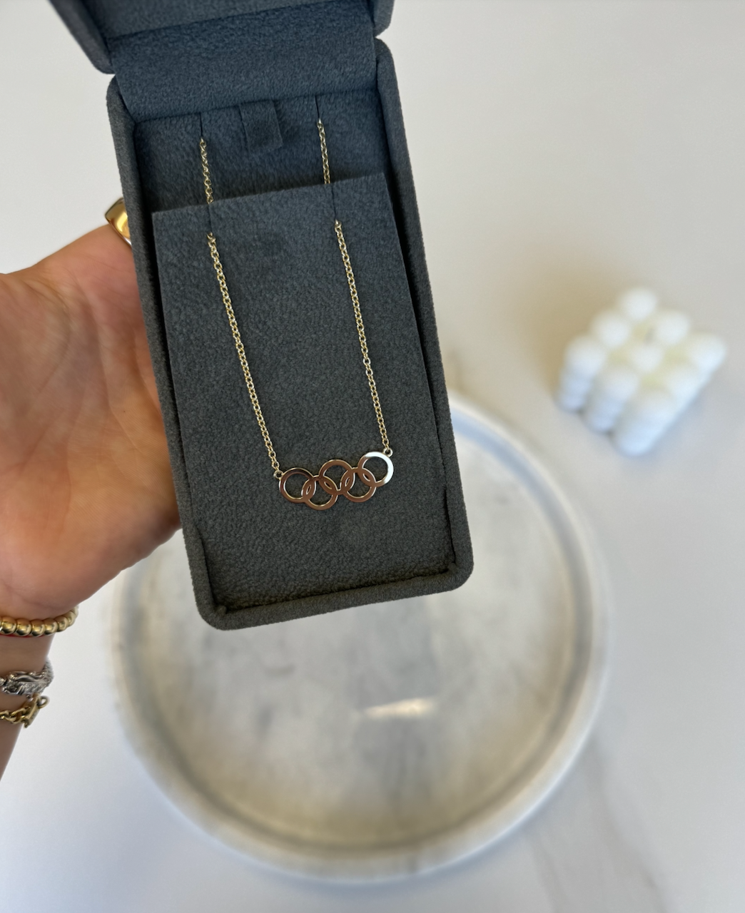 A hand holds an open gray jewelry box displaying the Gold Rings Necklace, featuring four interlocking gold rings—custom jewelry by Janet Heller. A blurred round tray and stacked white objects are visible in the background.