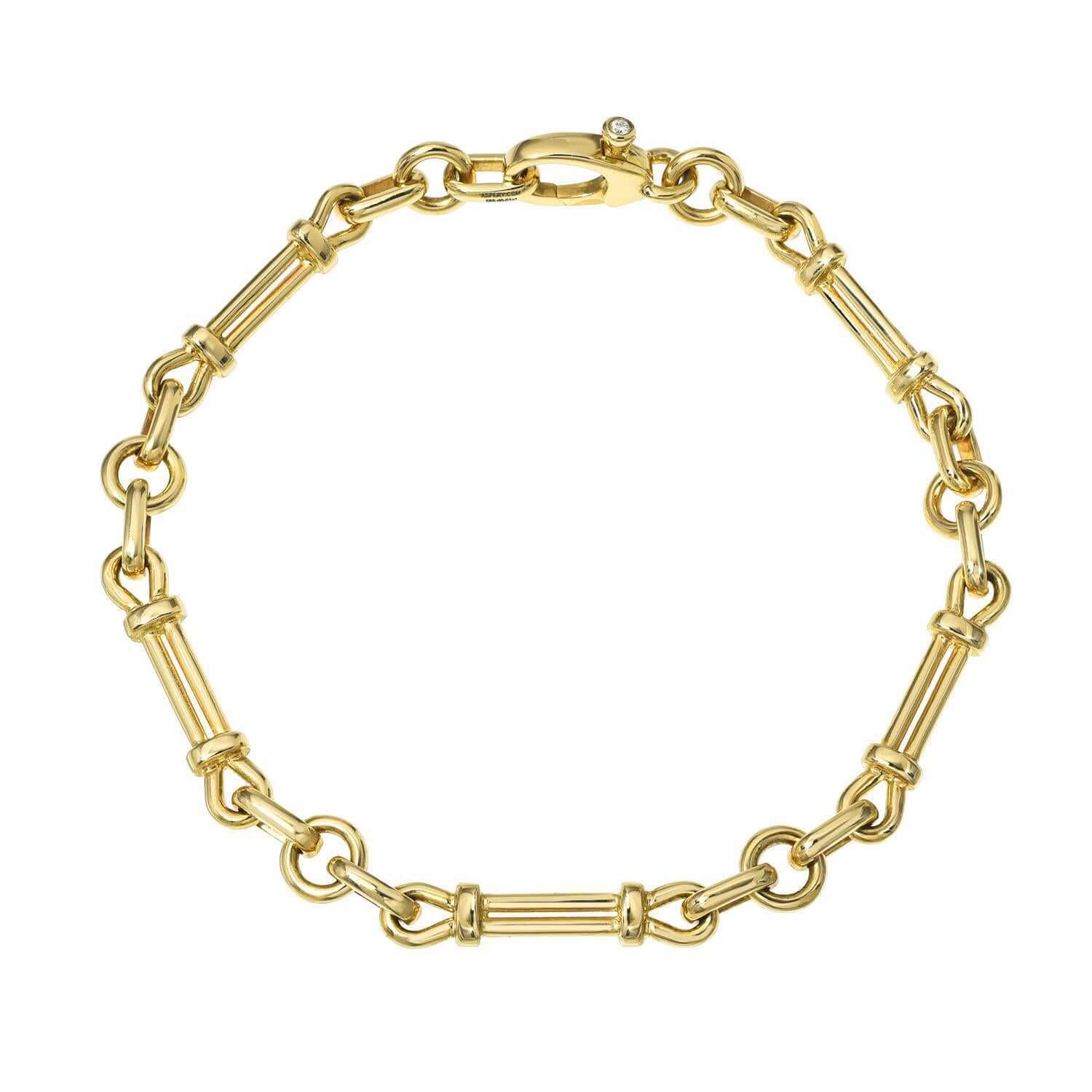 The Lisbon Bracelet by Janet Heller Fine Jewelry is a gold chain-link design with small loops and elongated cylindrical segments. It features a clasp adorned with a small diamond accent. The bracelet can be worn on its own or stacked with other bracelets.