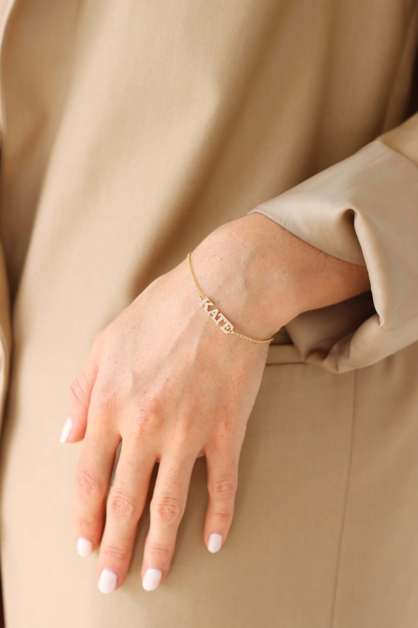 A close-up of a person's hand wearing the Test - Diamond Block Name Bracelet, featuring letter charms that spell KATE, against a beige outfit background.