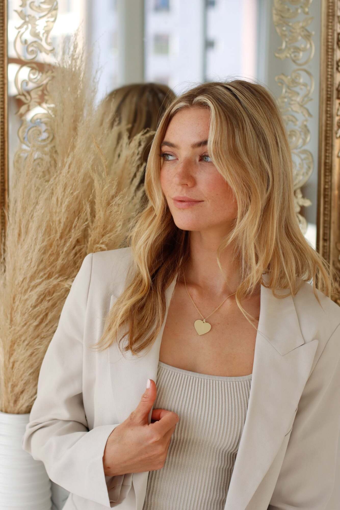 A woman with long blonde hair wearing a beige blazer poses indoors near a gold-framed mirror and pampas grass in a white vase, her Large Heart Engravable Pendant Necklace elegantly displaying an engraved message for a truly personalized keepsake.