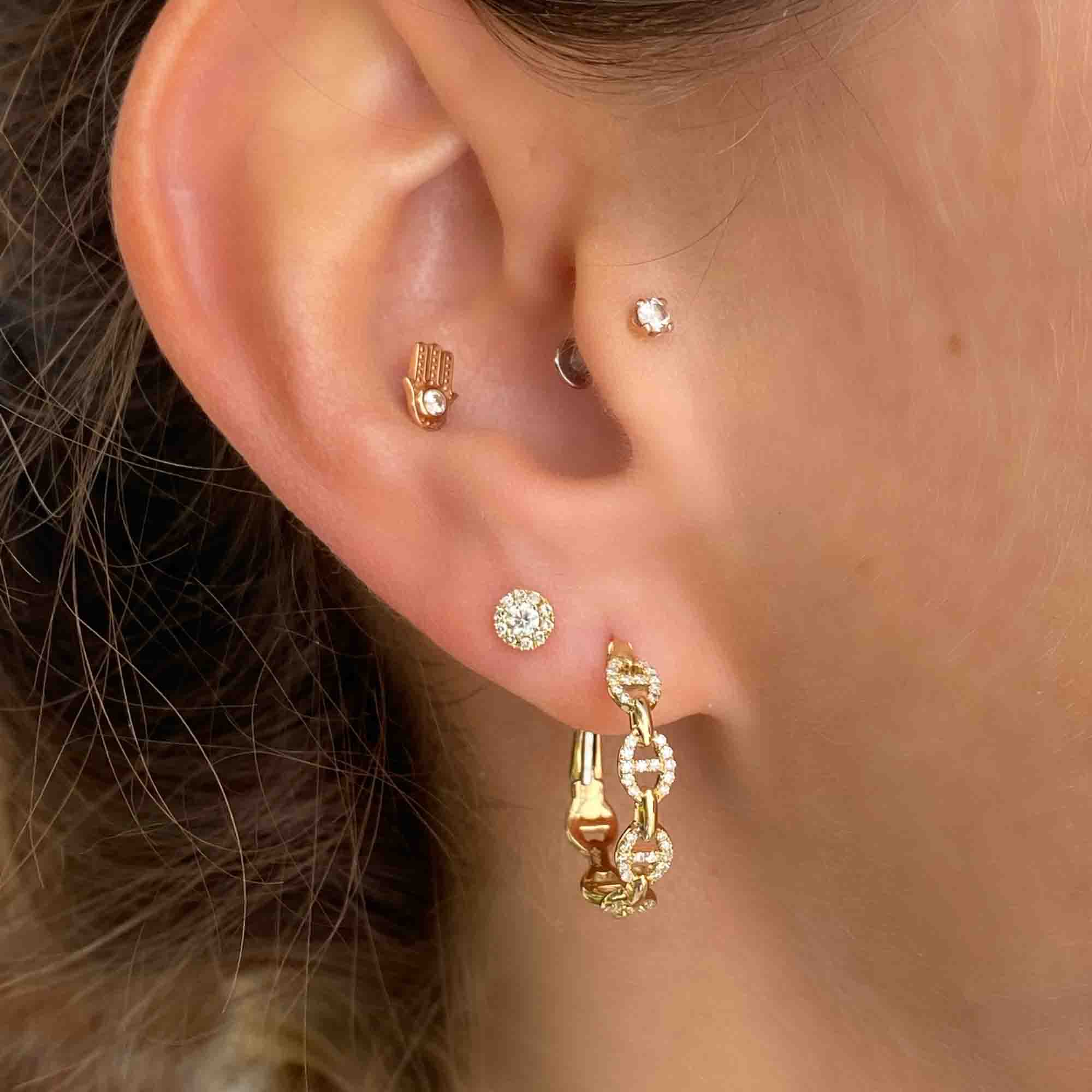 An ear displays stunning jewelry: gold earrings include a small stud, a tiny hoop, a diamond stud, and Diamond Matilda Hoops with chain design. Subtle skin is visible in the background.
