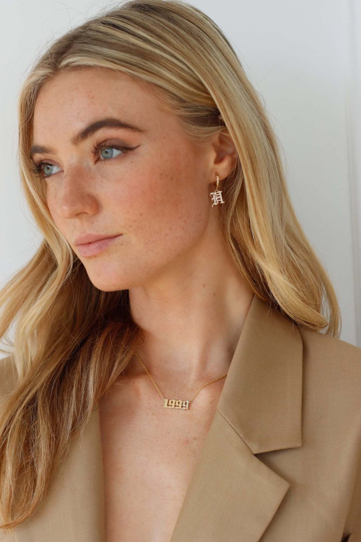 A woman with long blonde hair wears a tan blazer, gold earrings, and the Diamond Year Necklace—a gold personalized necklace featuring the numbers 1997. She looks to the side with a neutral expression against a plain white background.