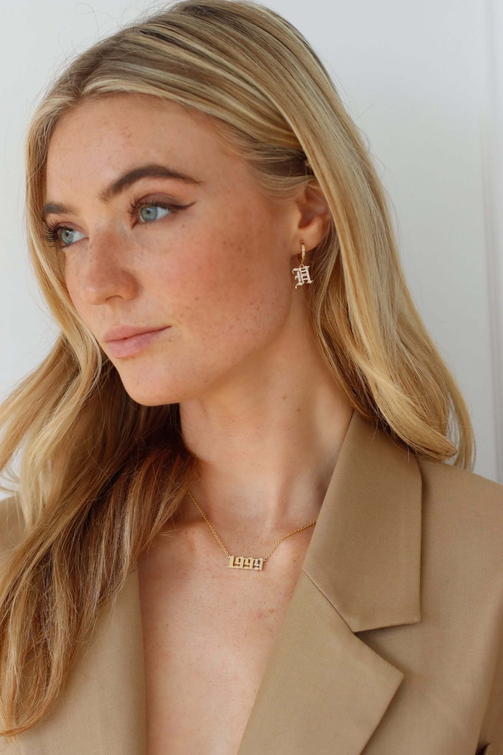 A woman with long blonde hair wears a tan blazer, gold huggie earrings from the Small Old English Initial Huggies collection, and a gold necklace with the numbers 1997. She looks to the side against a plain light background.