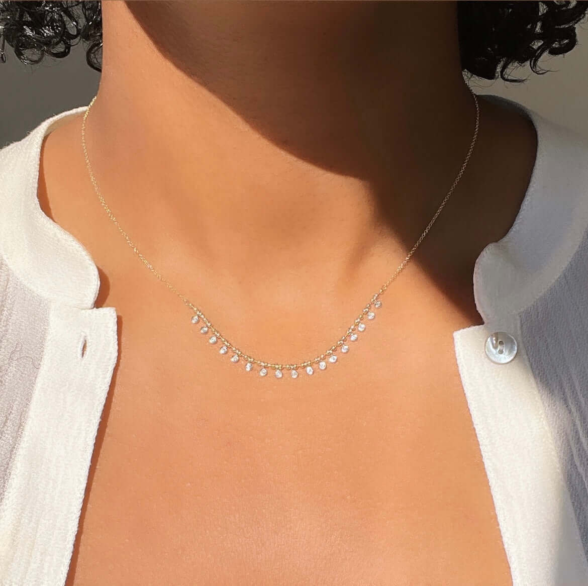 A person wears the Beaded Two Tone Diamond Necklace from Janet Heller Fine Jewelry. It rests delicately against their skin above a white, partially buttoned top. Soft lighting highlights the jewelry and clothing.