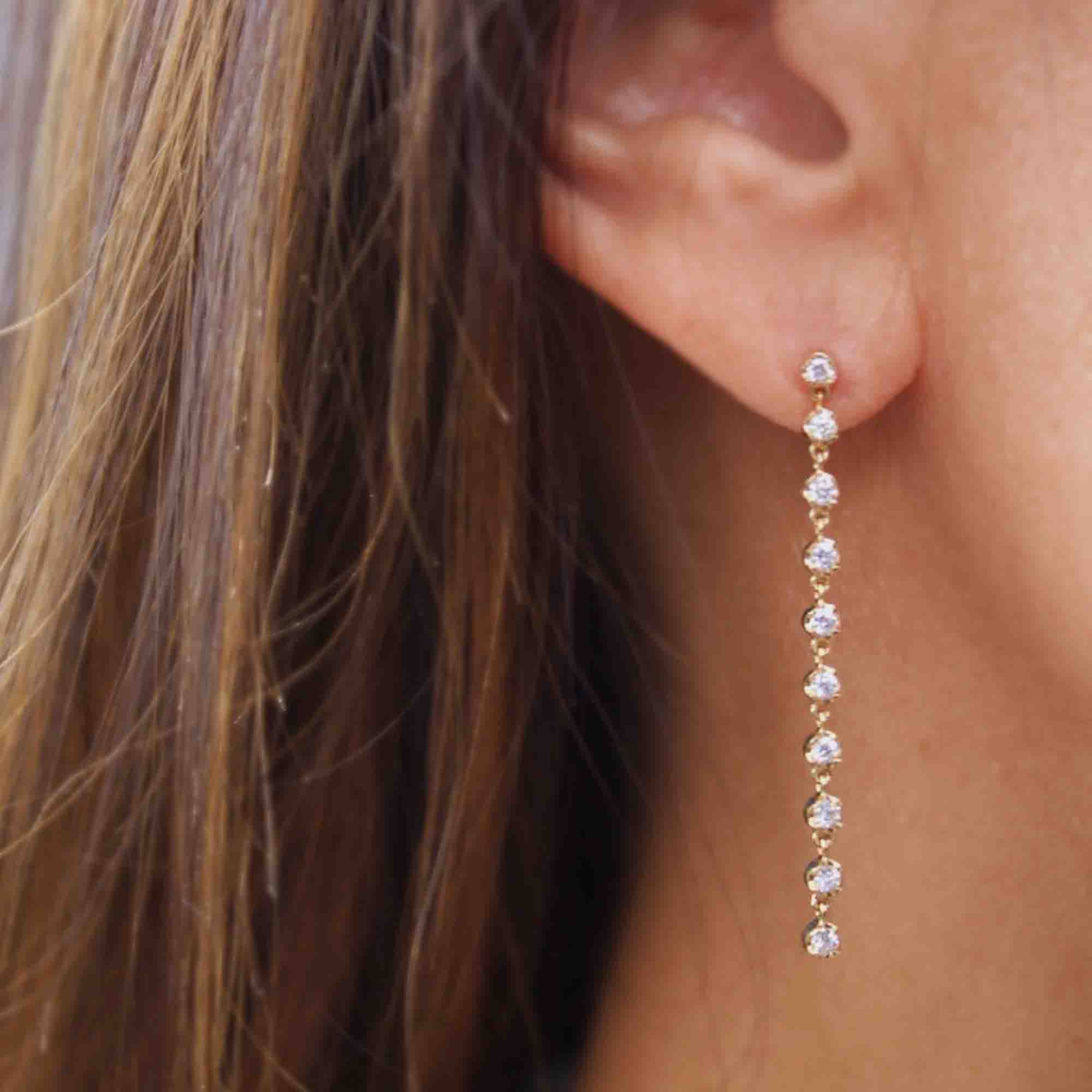 Close-up of a person wearing Janet Hellers 10 Drop Diamond Reagan Earrings, showcasing long, dangling fine jewelry with a series of small, round gemstones set in a straight line against the backdrop of the persons brown hair.