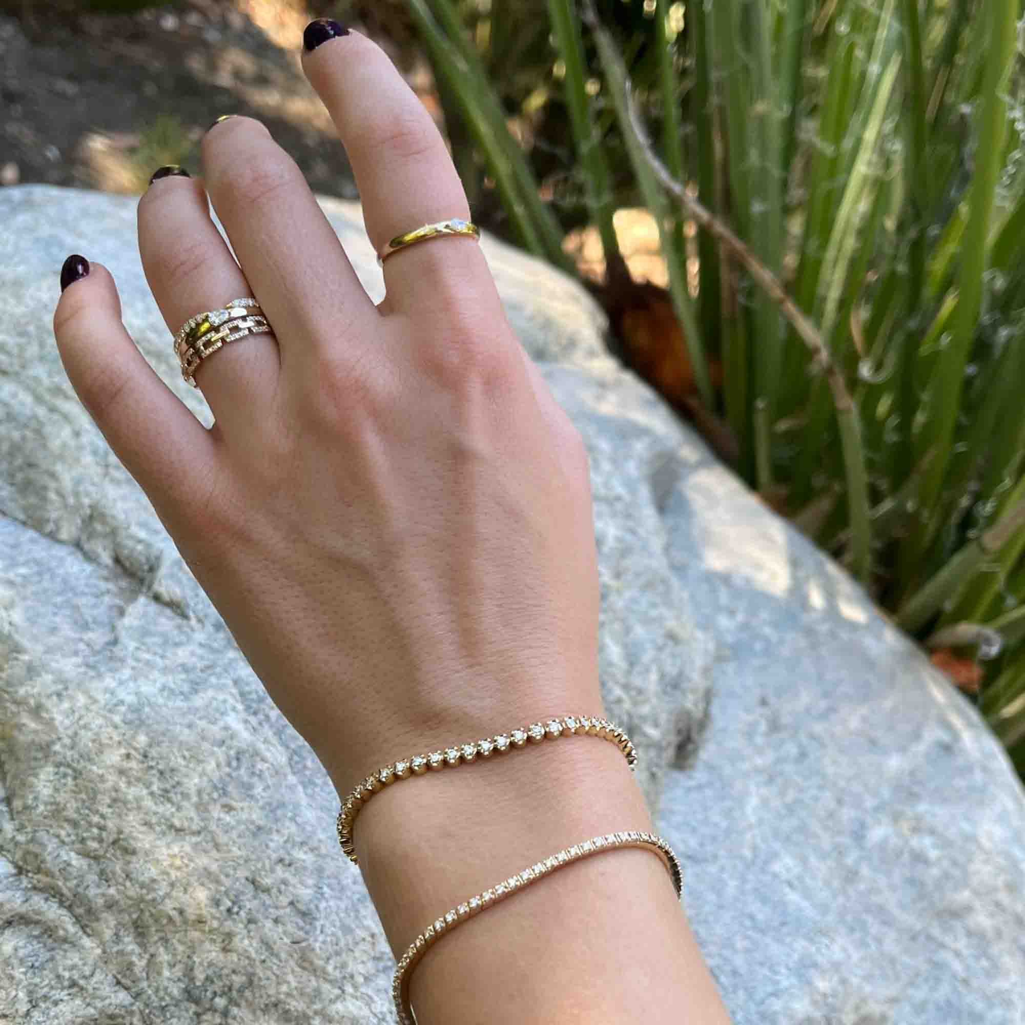A hand with dark nail polish wears gold rings and two Diamond Augustus Tennis Bracelets, each adorned with small diamonds, resting on a rock with green plants in the background.