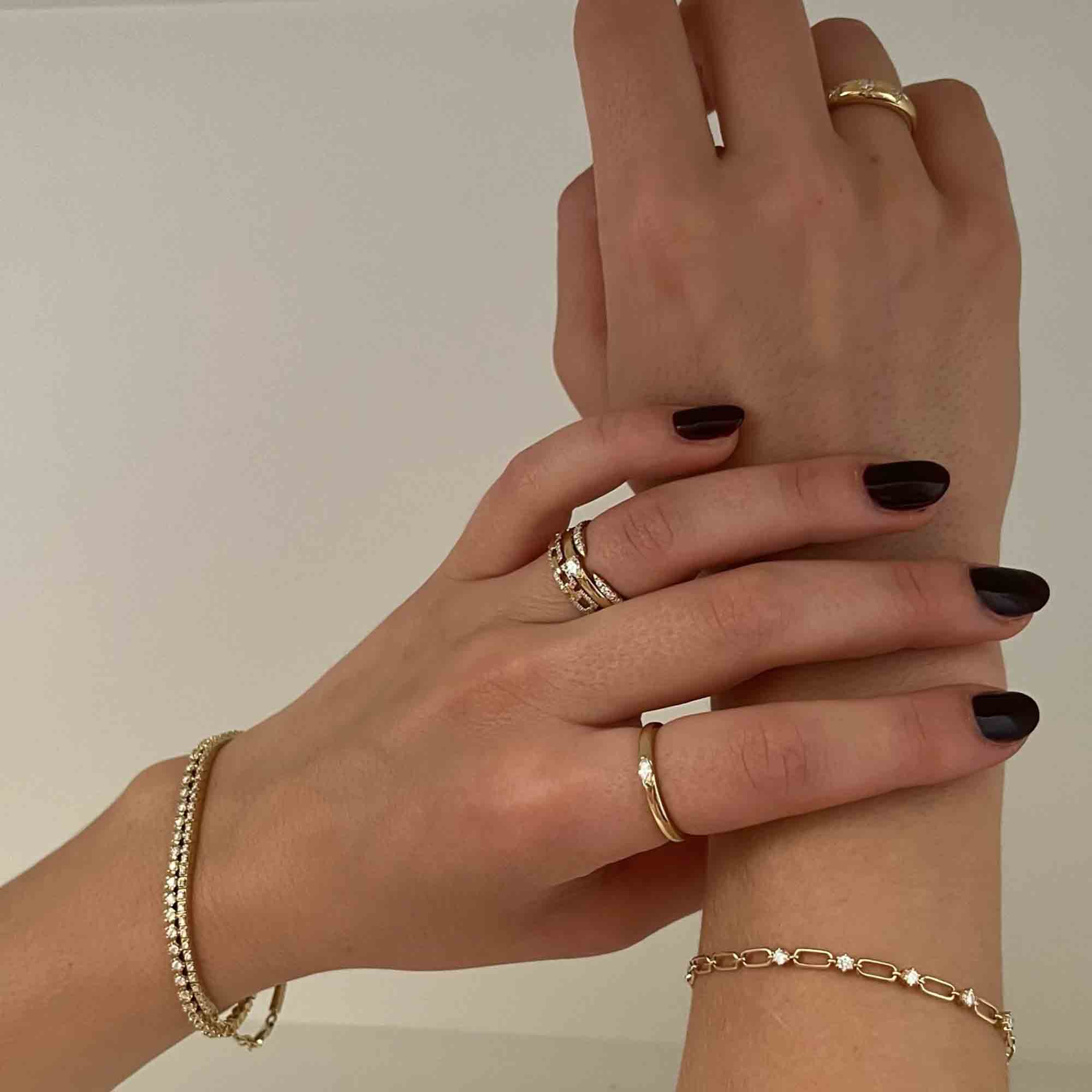 Two hands with dark nail polish display gold rings, a diamond ring, a gold chain bracelet, and the Diamond Augustus Tennis Bracelet against a neutral background.