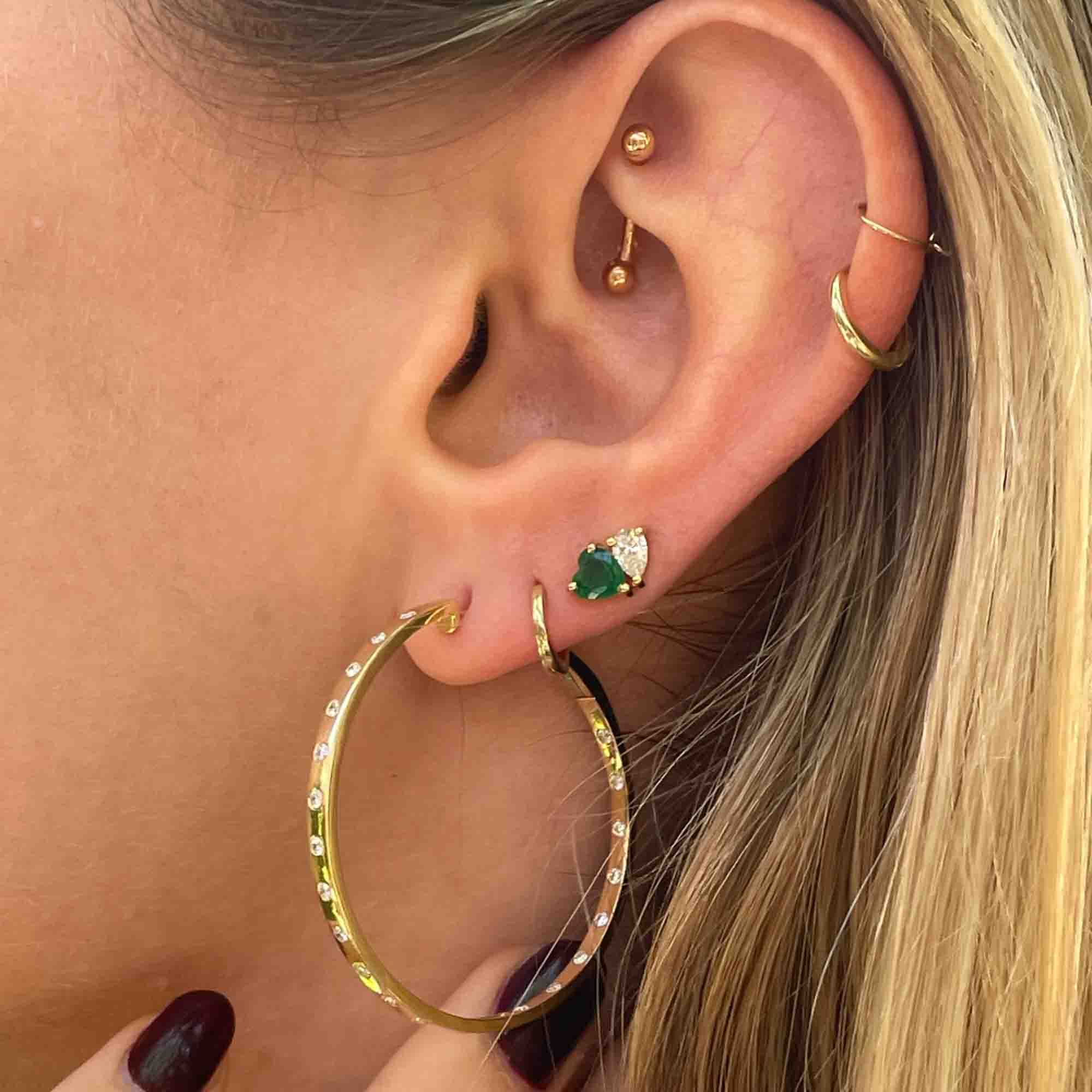 Close-up of an ear adorned with Janet Heller Fine Jewelry: Diamond Avery Hoops, a small hoop, a gem-accented green stud, plus helix hoop and snug stud. The blonde flaunts dark red nail polish, epitomizing elegance.