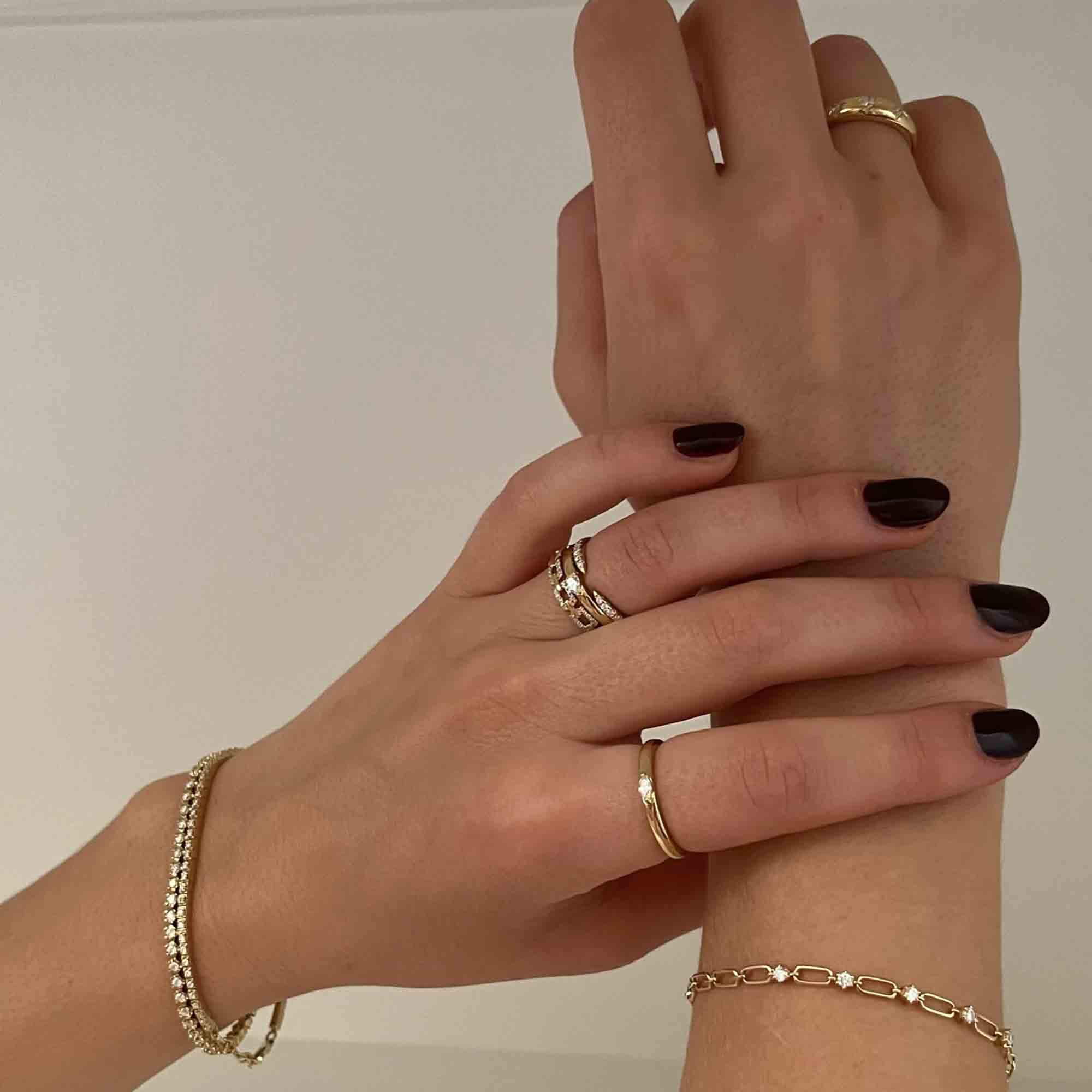Close-up of two hands adorned with custom jewelry: one hand features two rings and the Diamond Blakely Tennis Bracelet with dark manicured nails, while the other displays a ring and delicate chain bracelet against a plain, light background.