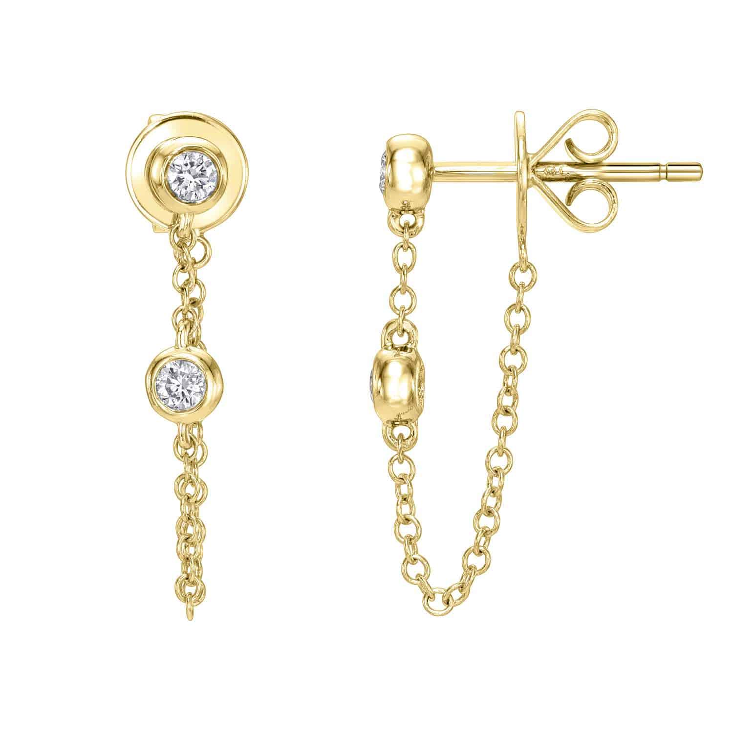 The Double Bezel Diamond Studs by Janet Heller Fine Jewelry are crafted in 14KT gold. The earrings feature dangling chains, each showcasing two sparkling round diamonds set in a gold bezel. The studs can be worn alone or layered along the ear with other earrings.