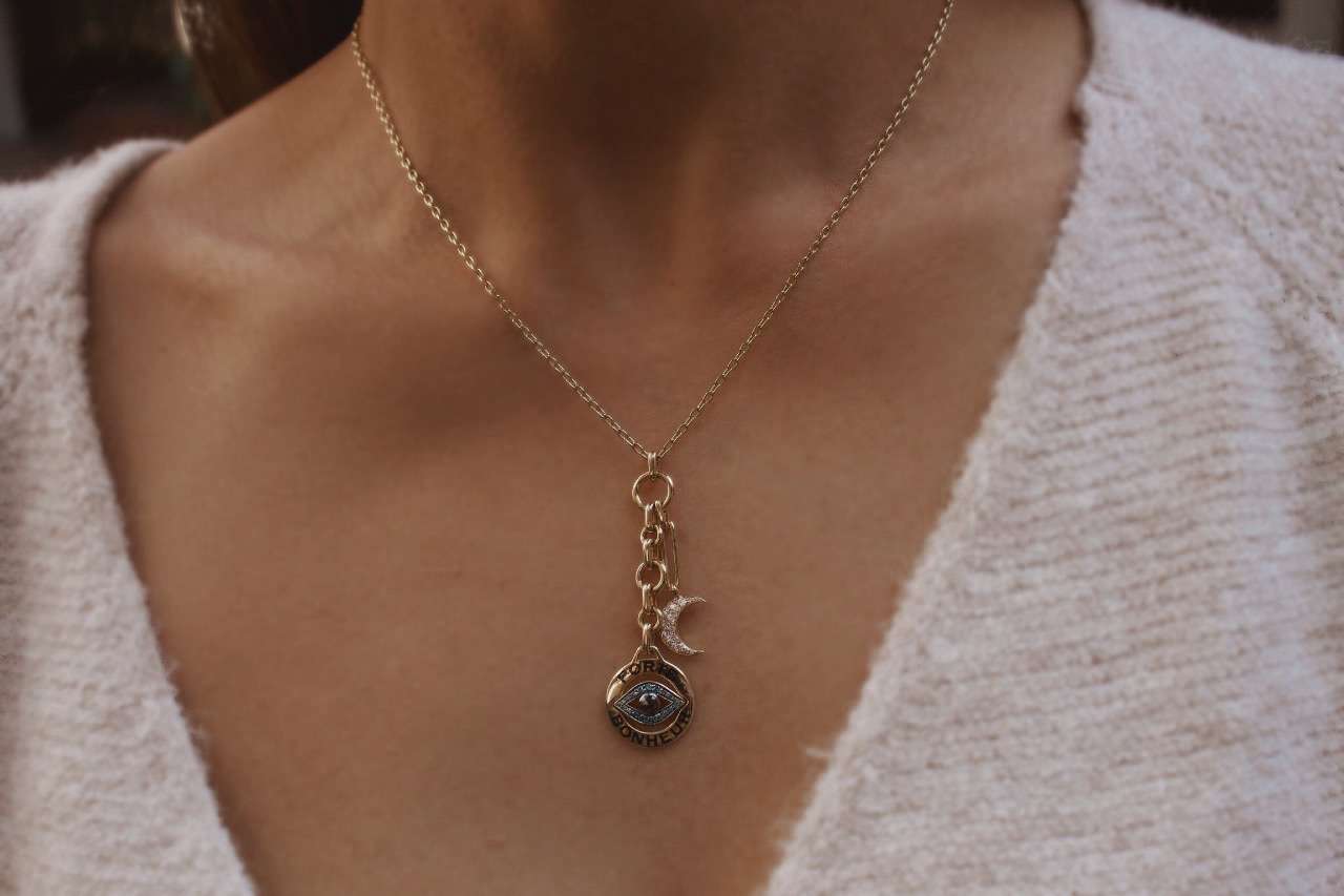 Close-up of a person wearing the Small Diamond Porte Bonheur Necklace, featuring an eye pendant and crescent moon charm. They are dressed in a soft-textured, light-colored top that highlights this exquisite piece of jewelry.