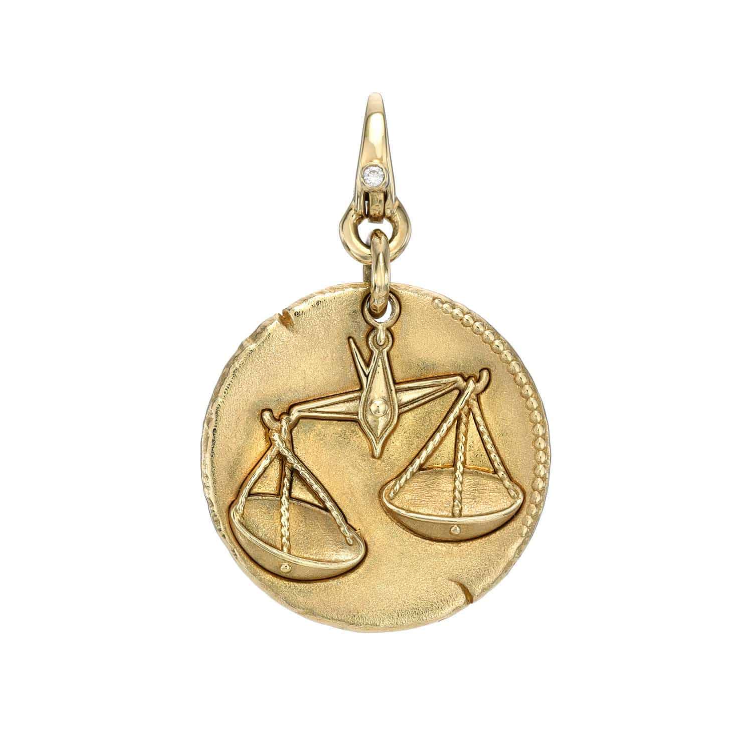 The Zodiac Sign Charm is a round gold pendant featuring a raised balanced scale design representing Libra. It has a textured edge and a small loop at the top for attaching to a chain.