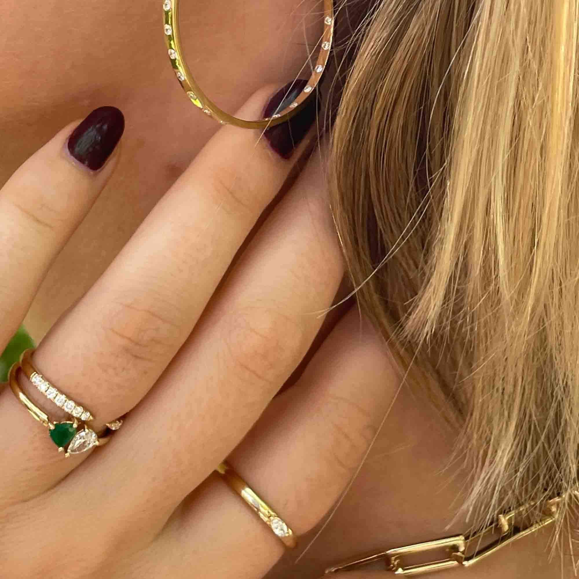 Close-up of a person showcasing fine jewelry. Theyre wearing a large gold hoop earring and the Emerald & Diamond Toi et Moi Ring with a green gemstone and diamonds. Their nails are painted dark, and they have blonde hair—ideal pieces to preorder for your collection.
