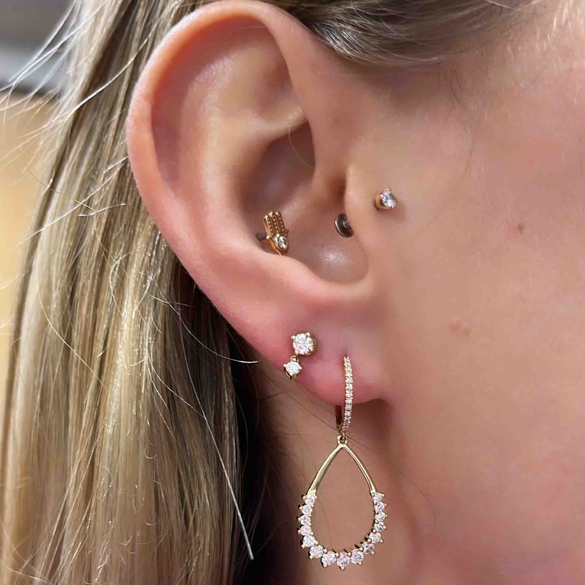 Close-up of an ear with fine jewelry: Open Pear Diamond Earring, sparkling teardrop-shaped, graces the lobe. A small stud above and a stone-accented hoop on the helix, plus a simple stud on the tragus. For customizations, contact inquiries@janethellerfinejewelry.com. Hair elegantly tucked back.