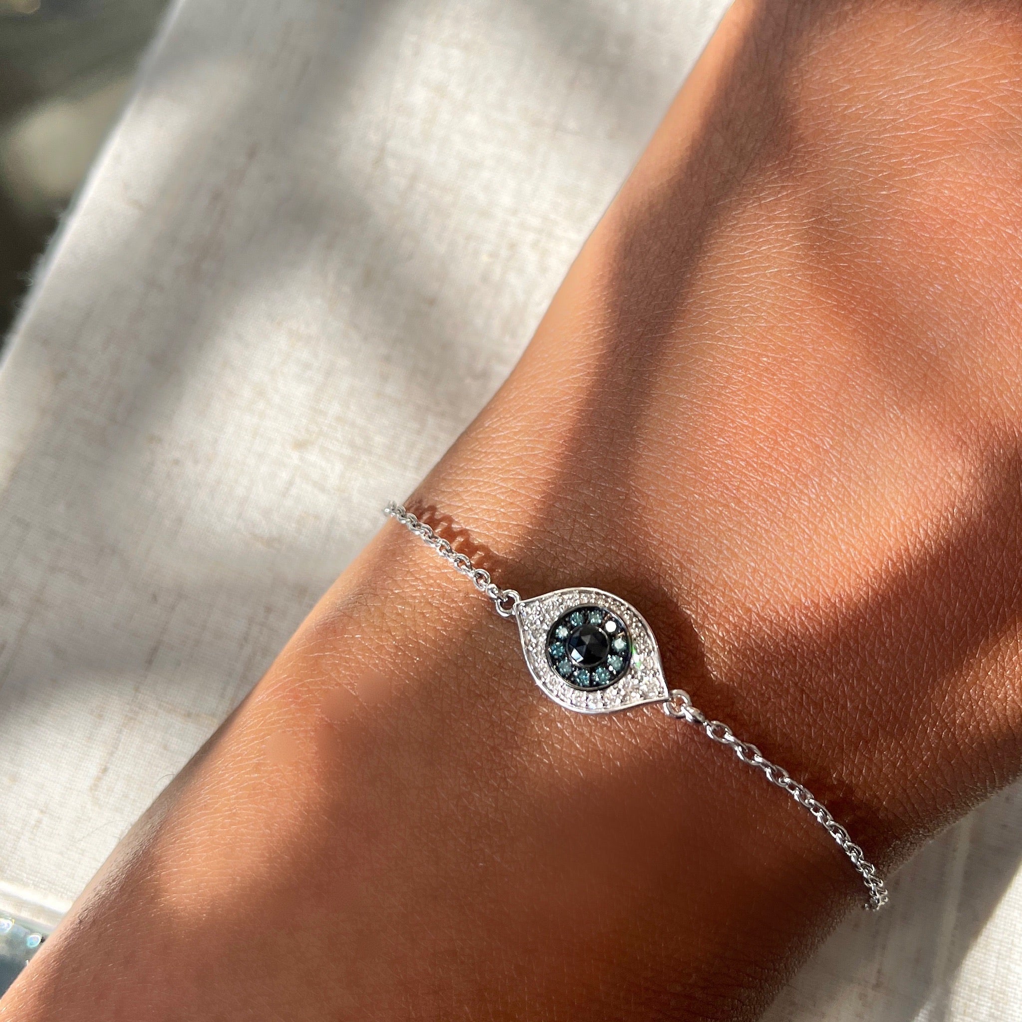 A hand wearing the Blue Diamond Evil Eye Bracelet from janethellerfinejewelry, featuring an eye-shaped charm with a central black gemstone surrounded by smaller clear stones, rests on a textured light surface where soft shadows form a subtle pattern.