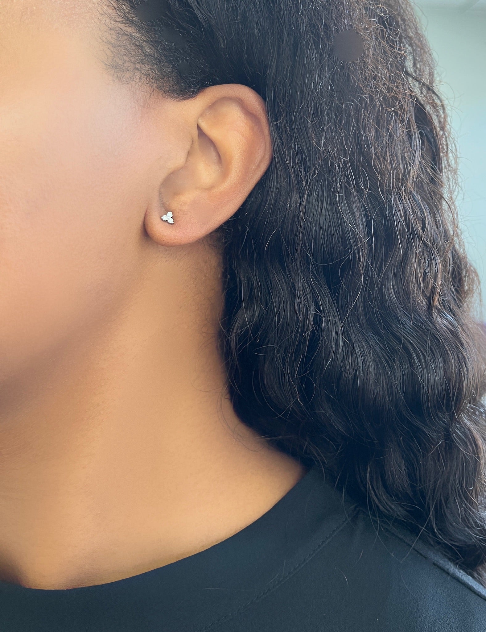 The close-up captures a Cluster Diamond Trio Stud from janethellerfinejewelry resting on someones left ear. With long, wavy dark hair and a dark top, the shot elegantly highlights the beauty of fine jewelry against a softly blurred background.