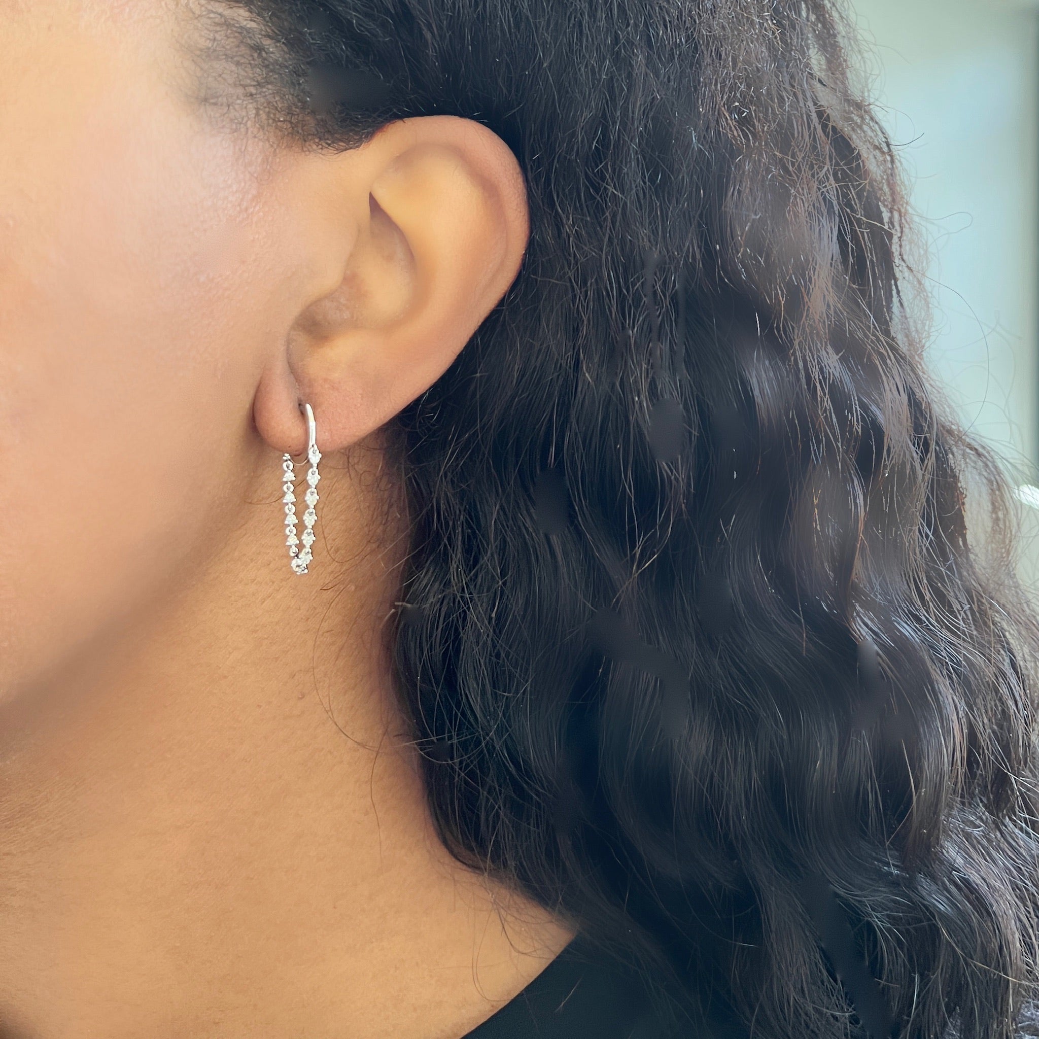 A close-up shot showcases a woman's ear adorned with the Diamond Cascade Drop Huggies from Janet Heller Fine Jewelry—sleek silver hoops elegantly set with sparkling stones. Her long, curly dark hair and medium skin tone beautifully complement the fine jewelry, while the softly blurred background accentuates its refined charm.