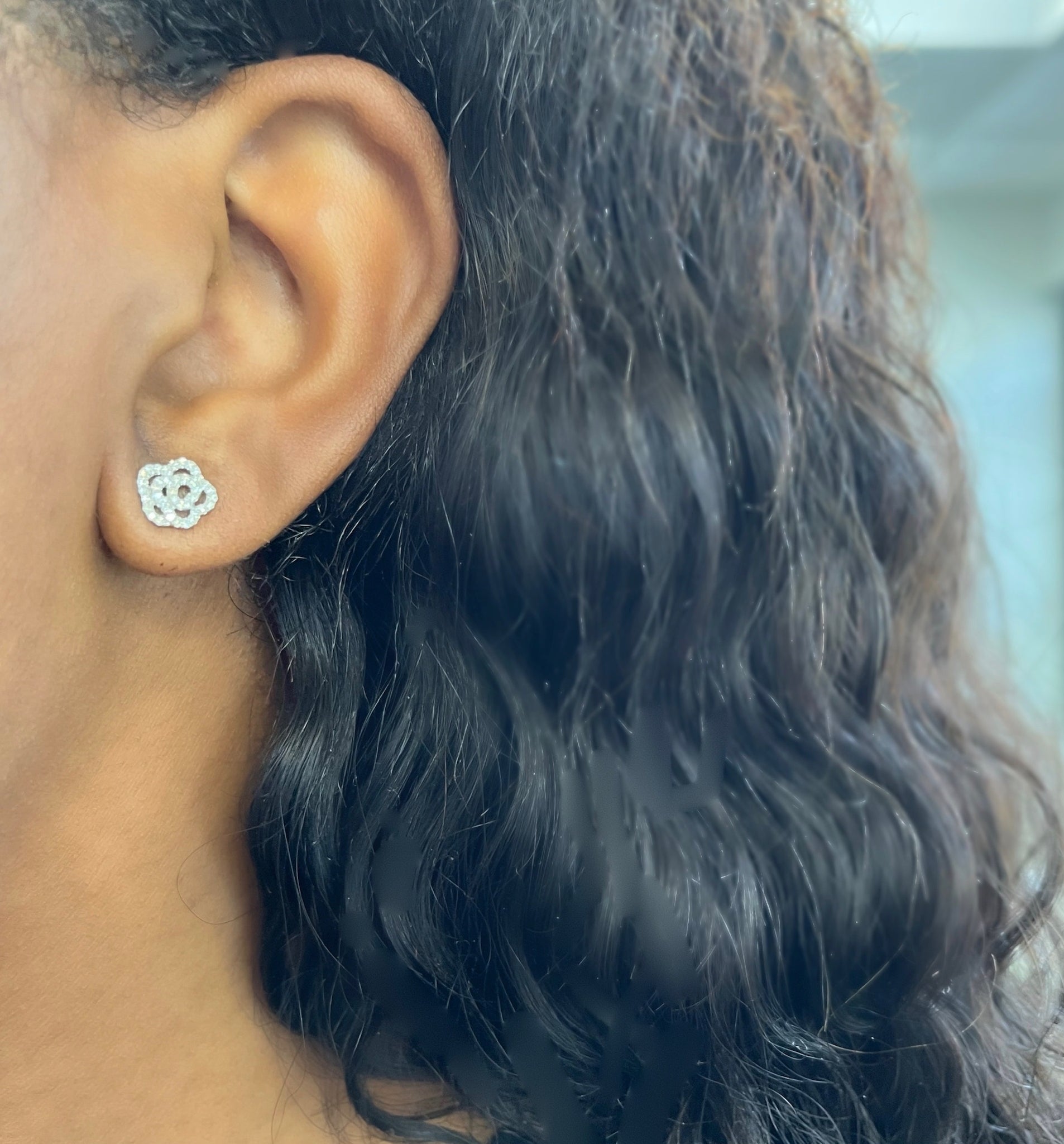 A close-up of a person’s ear with long, dark, wavy hair, wearing the Diamond Rose Studs—a small, silver, flower-shaped stud earring from Janet Heller Fine Jewelry. Perfect for those seeking custom jewelry or with specific jewelry inquiries. Price: $600.00.