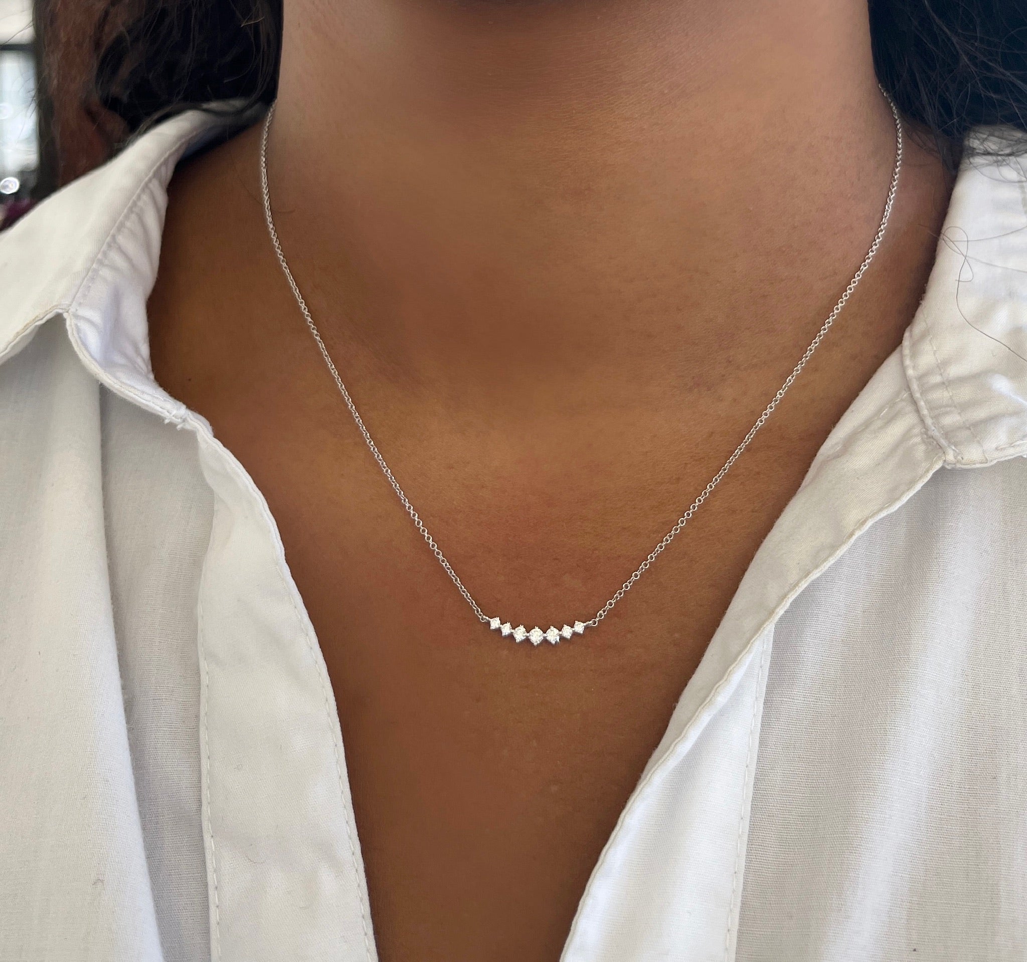 A person wearing a white collared shirt and a delicate silver necklace featuring graduating U-shaped diamond charms arranged in a row—a timeless piece from Janet Heller Fine Jewelry’s 'Graduating U Shaped Diamond Necklace' (NEC-1377-W), celebrated for its exquisite custom and elegant fine jewelry collections.