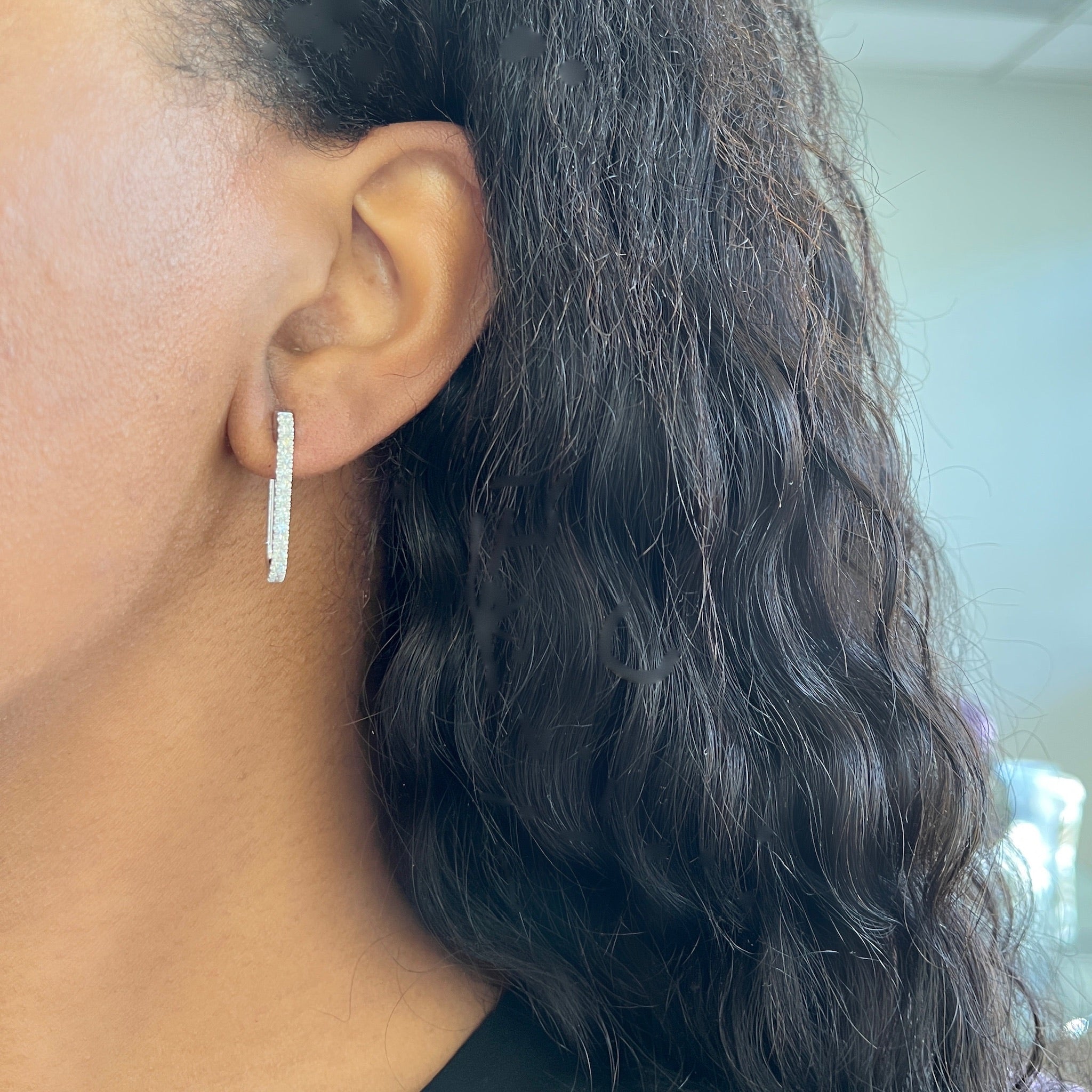 A close-up of a woman's ear adorned with the Mini Diamond Alice Earrings—a sparkling, vertical rectangular hoop that adds an elegant touch to her look. She has long, wavy dark hair, and the softly blurred background highlights the fine jewelry.