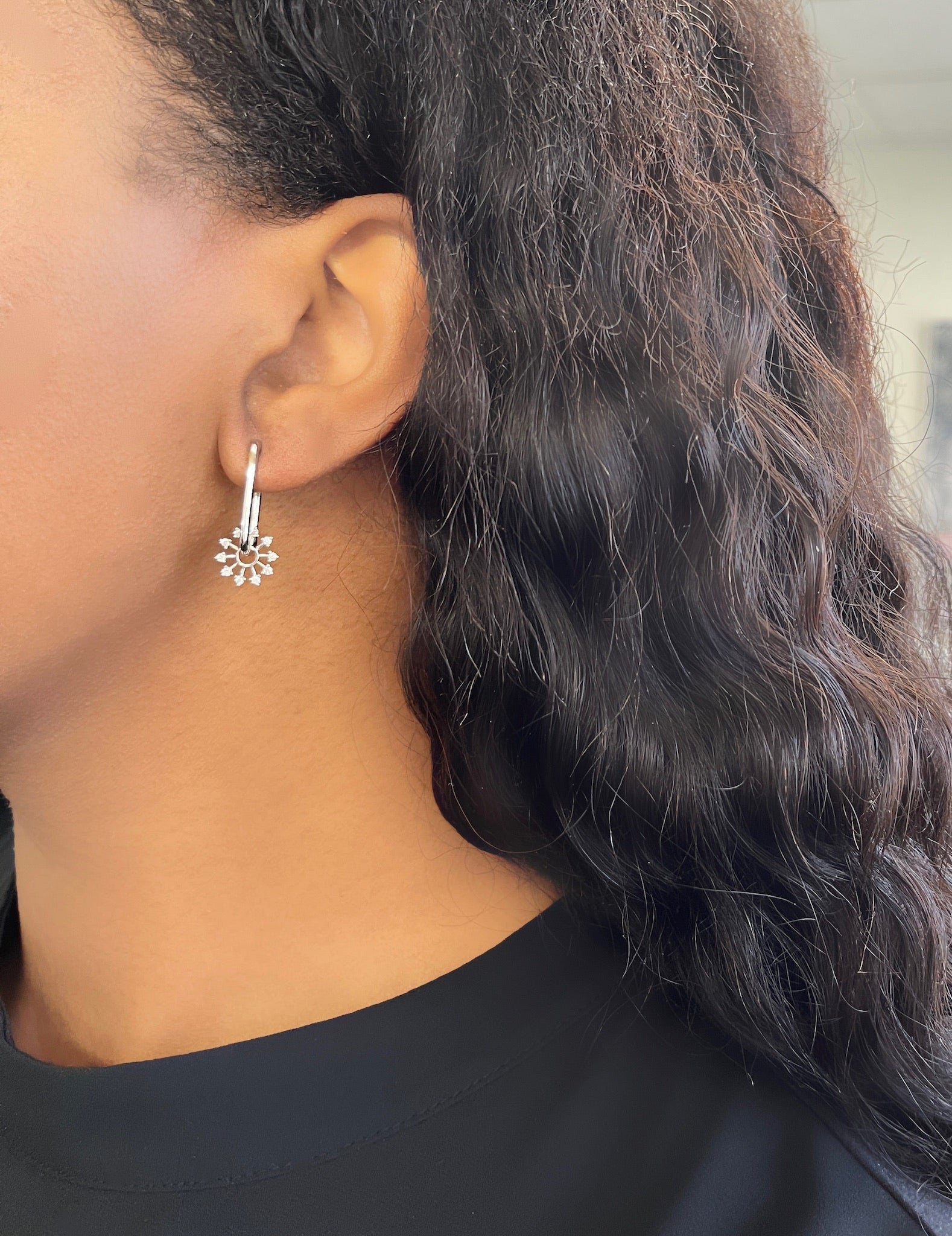 Close-up of a person’s left ear with wavy dark hair, wearing the Oval Diamond Star Charm Hoops from Janet Heller Fine Jewelry—a silver hoop earring featuring a dangling star-shaped charm. The person is dressed in a black top, highlighting how fine jewelry elevates everyday style.