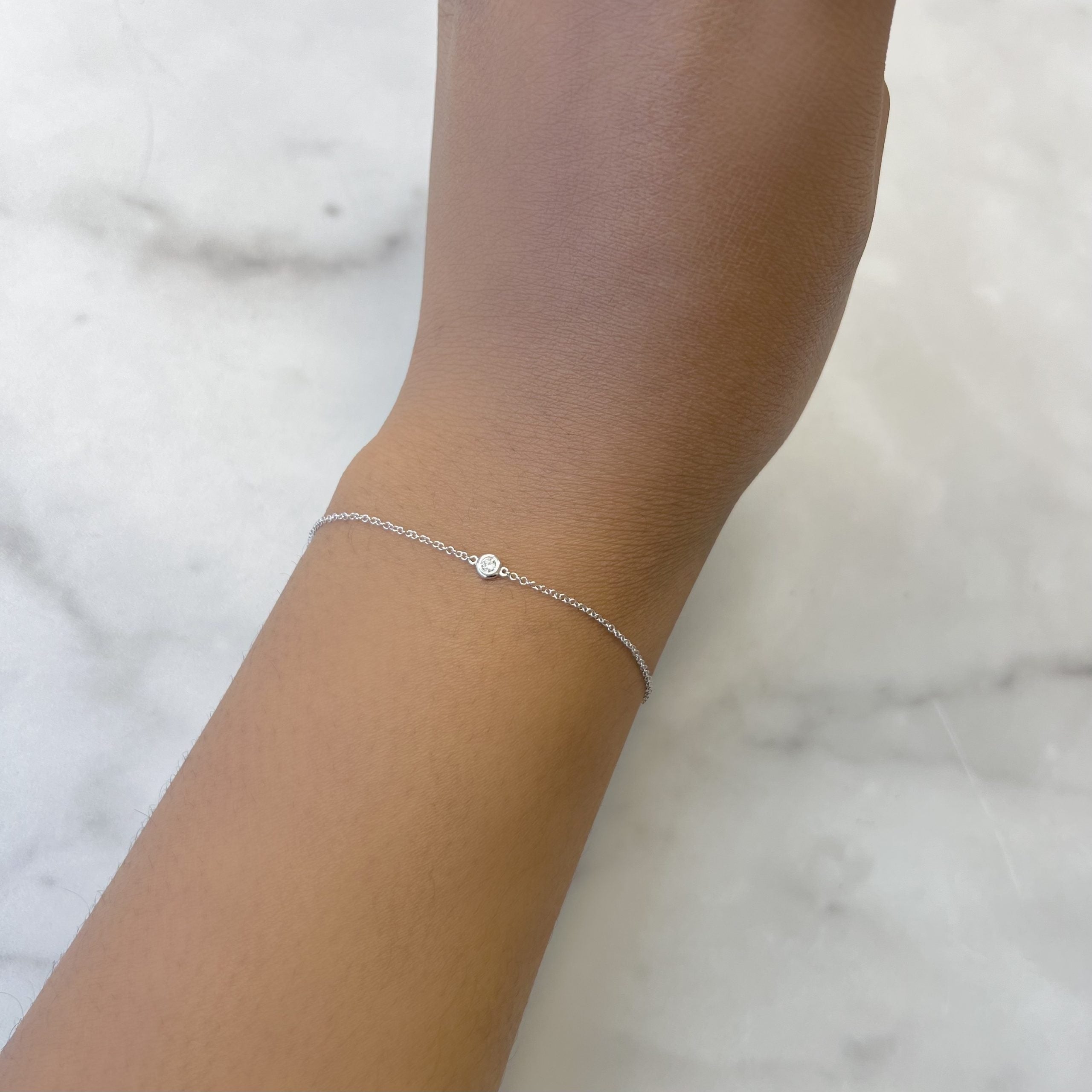 A hand wearing the Single Diamond Bezel Bracelet, a delicate fine jewelry piece with a circular charm, rests on a light marble surface. The thin, simple design offers endless customizations to suit your personal style.