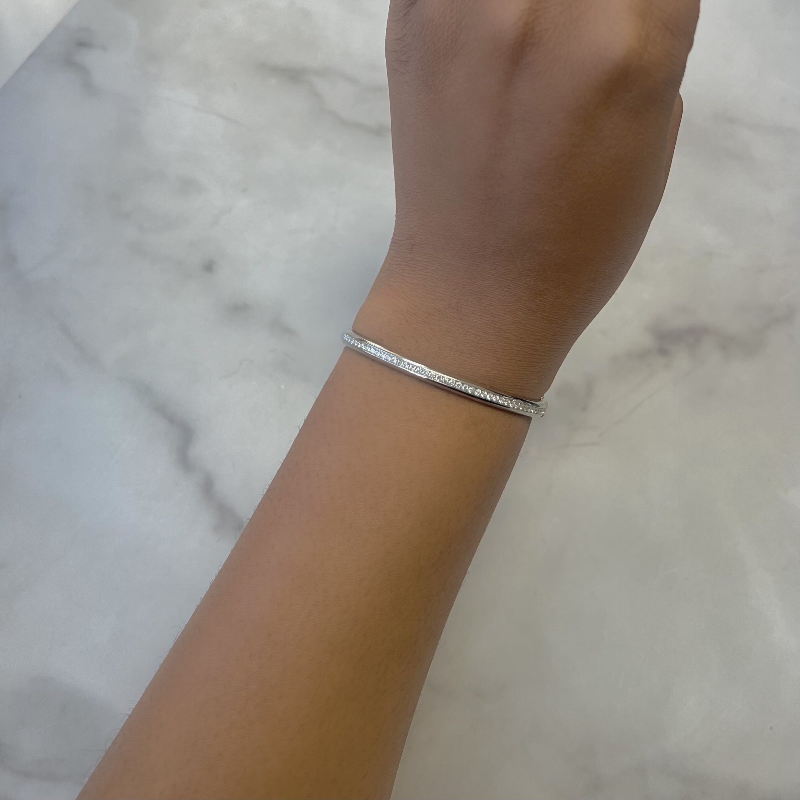 A close-up of a wrist features a Single Row Diamond Dome Bangle from janethellerfinejewelry against a light marble backdrop. The silver bracelets slight sheen complements the medium brown skin tone, with room for customizations to make it uniquely yours.