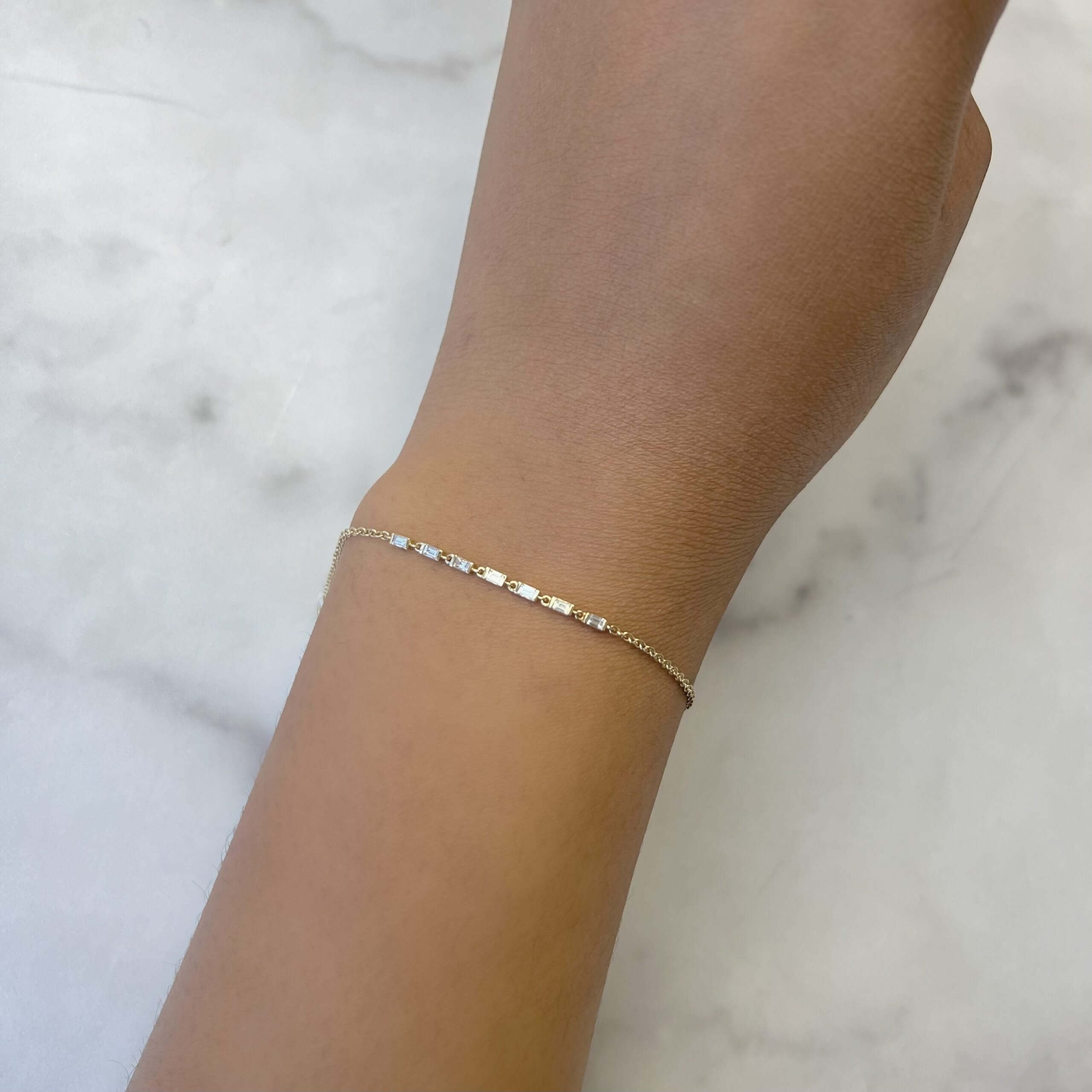 A close-up shows a wrist adorned with the Baguette Diamond Adda Bracelet from Janet Heller Fine Jewelry, featuring small, evenly spaced rectangular links. The backdrop is a soft, light-colored surface.