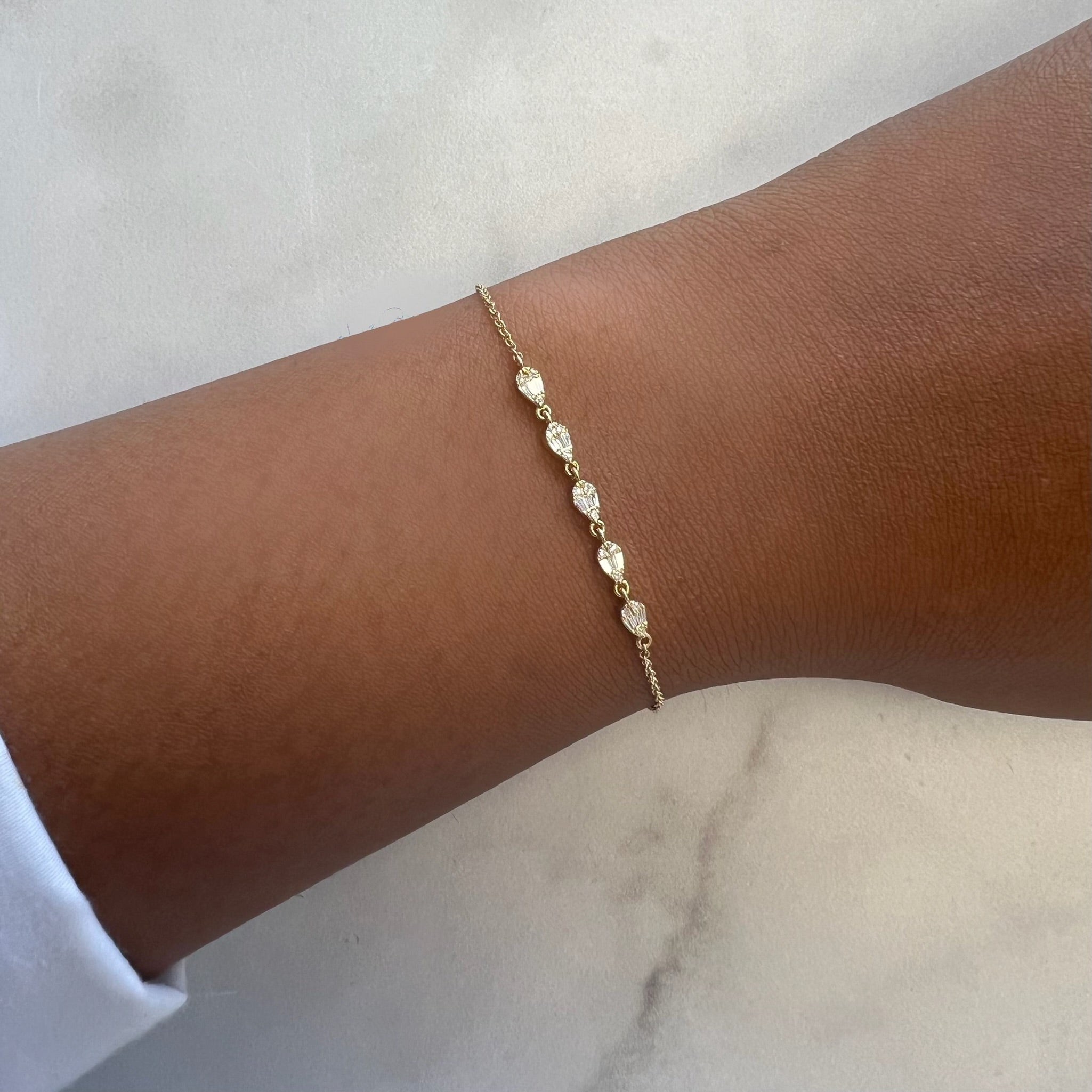 A close-up shows a wrist graced by the Baguette Diamond Pear Shaped Bracelet, featuring small teardrop-shaped charms. The background is a smooth, light-colored surface, complemented by a white sleeve adding elegance to the jewelry.