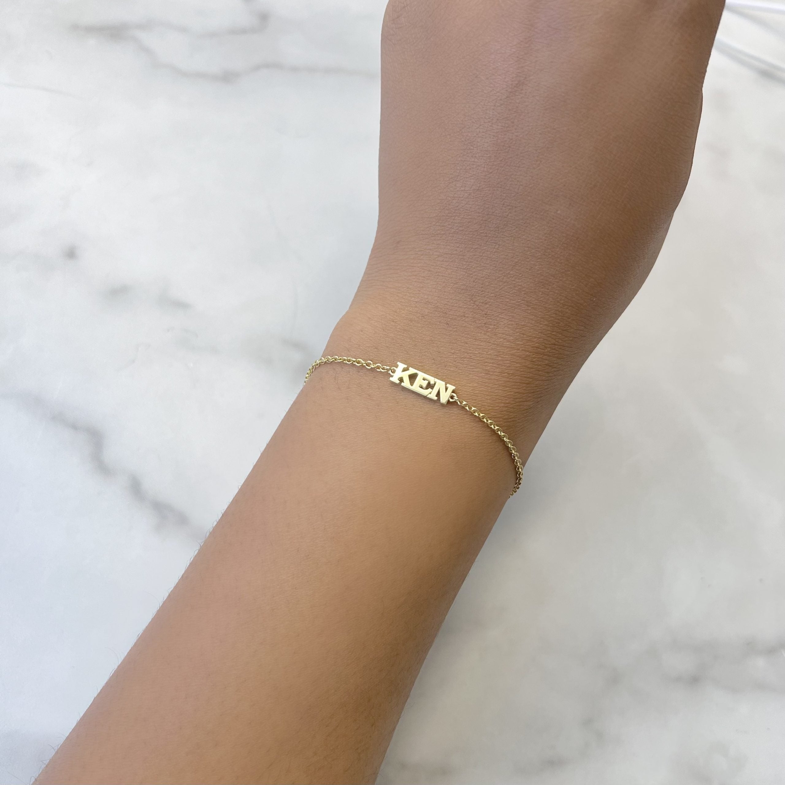 A close-up shows a wrist adorned with the Block Name Bracelet, a delicate gold piece featuring KEN on a thin chain. The bracelets uniqueness is accentuated by the light marble background.
