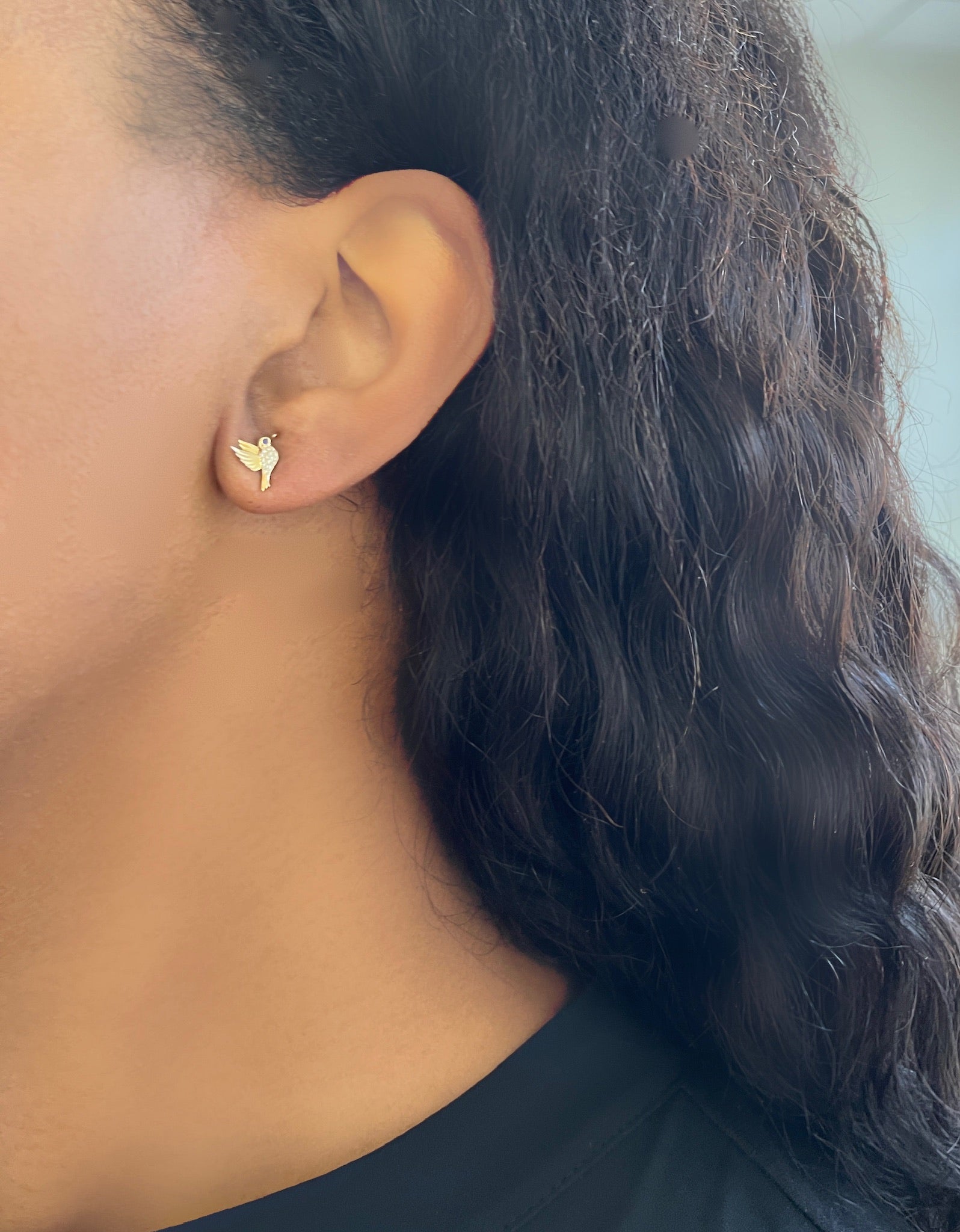 Close-up of a persons ear adorned with Blue Sapphire & Diamond Hummingbird Studs by Janet Heller Fine Jewelry. The person, with long, wavy dark hair, wears a black top against a softly blurred background.