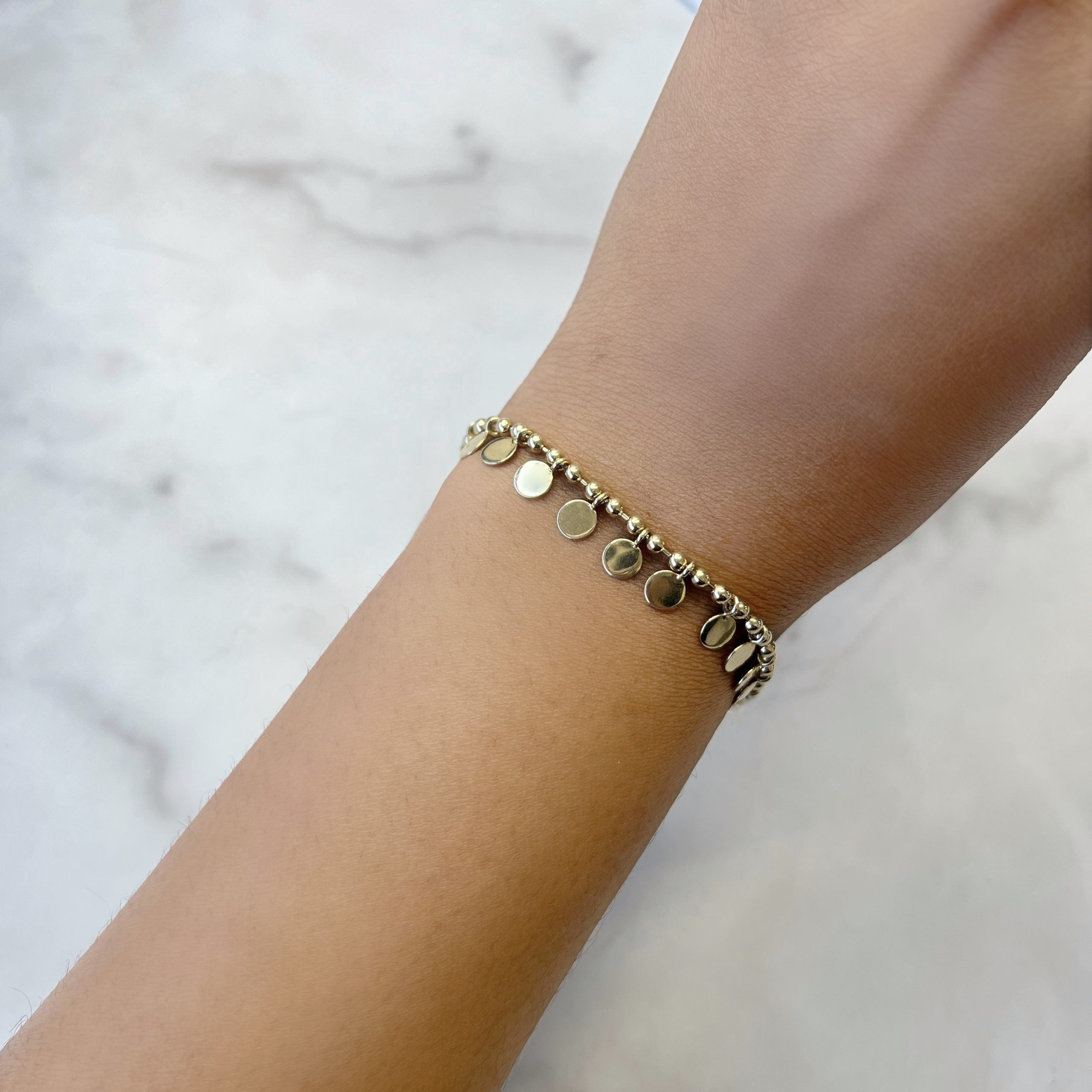 The elegantly adorned hand rests on a marble background, showcasing the refined beauty of fine jewelry with the Coin Bracelet, featuring small round charms in gold.