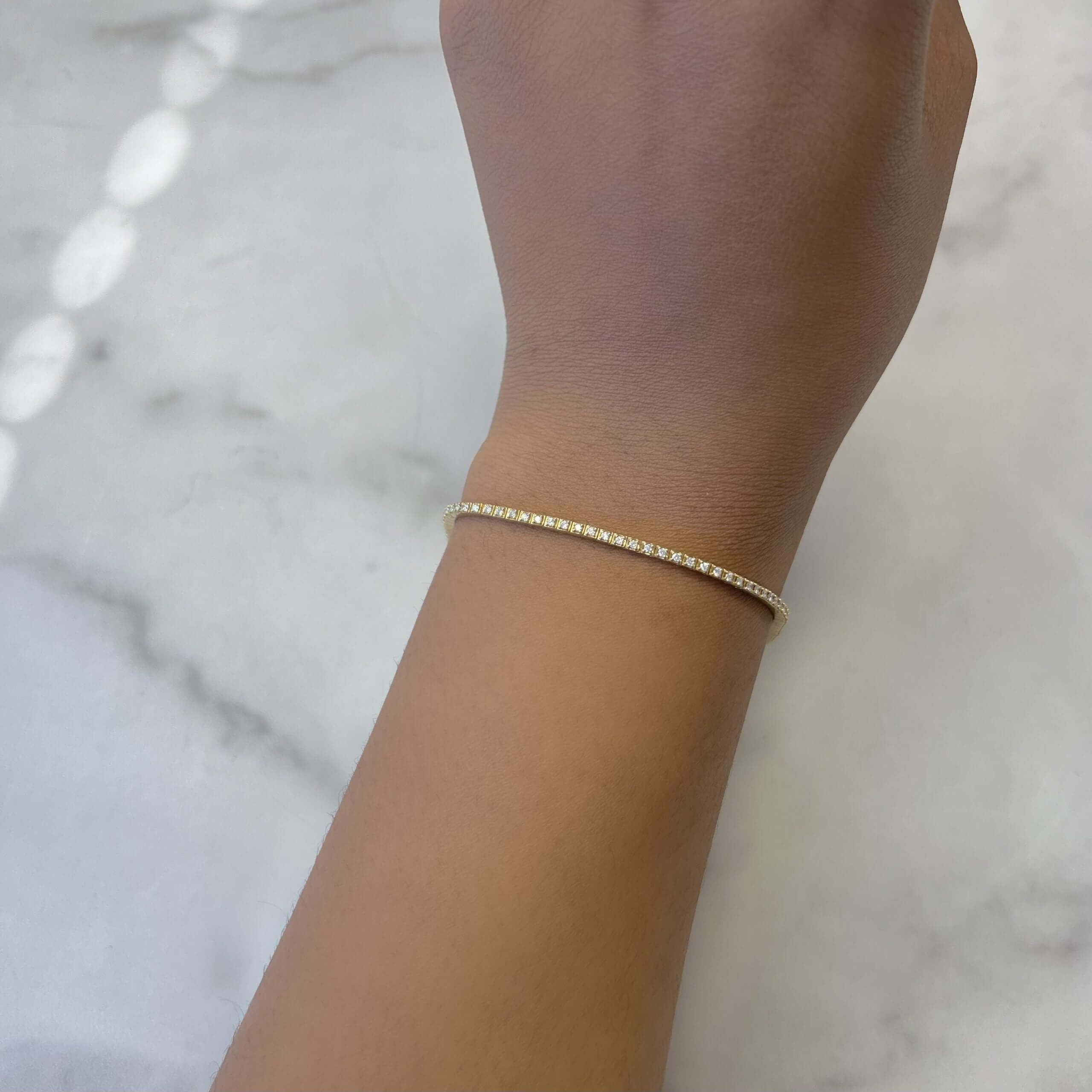 The Diamond Augustus Tennis Bracelet by Janet Heller Fine Jewelry elegantly adorns the wrist, its minimalist gold line resting on a light marble surface while tiny diamonds add a delicate sparkle.
