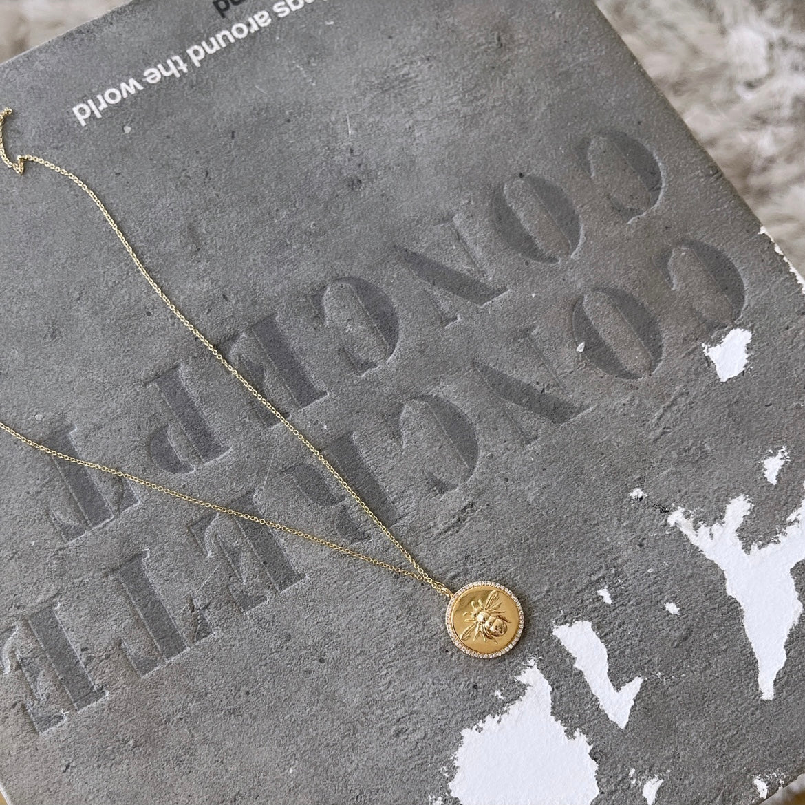 The Diamond Bee Pendant Necklace from Janet Heller Fine Jewelry, featuring a bee design on a round pendant, rests on a textured concrete surface showing incomplete white text reading CONC... RELATED.